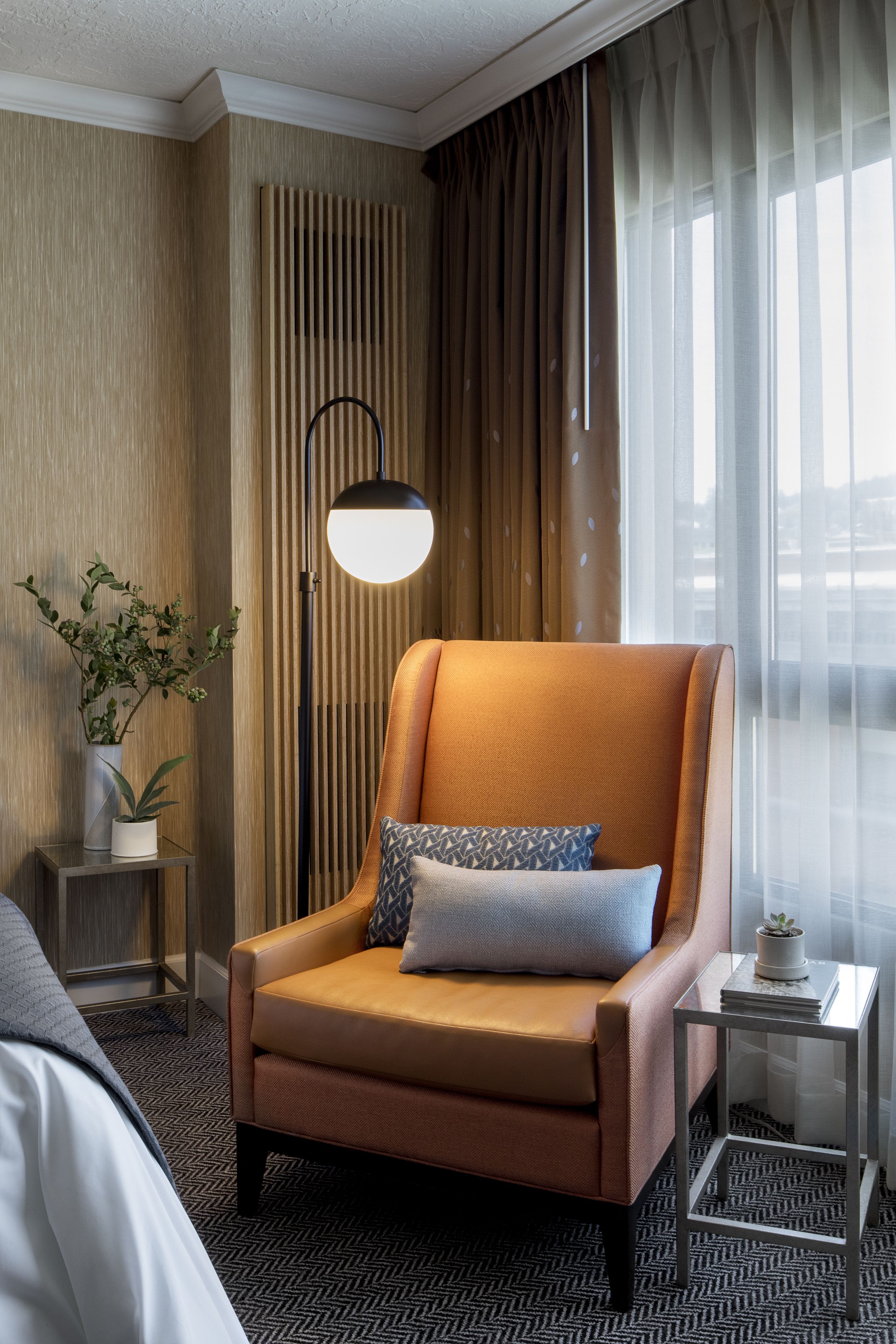 Habitación de hotel con un sillón de cuero, una mesita auxiliar con una lámpara, una planta en maceta y vistas a la ciudad a través de cortinas translúcidas.