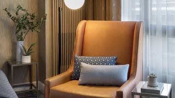 Habitación de hotel con un sillón de cuero, una mesita auxiliar con una lámpara, una planta en maceta y vistas a la ciudad a través de cortinas translúcidas.