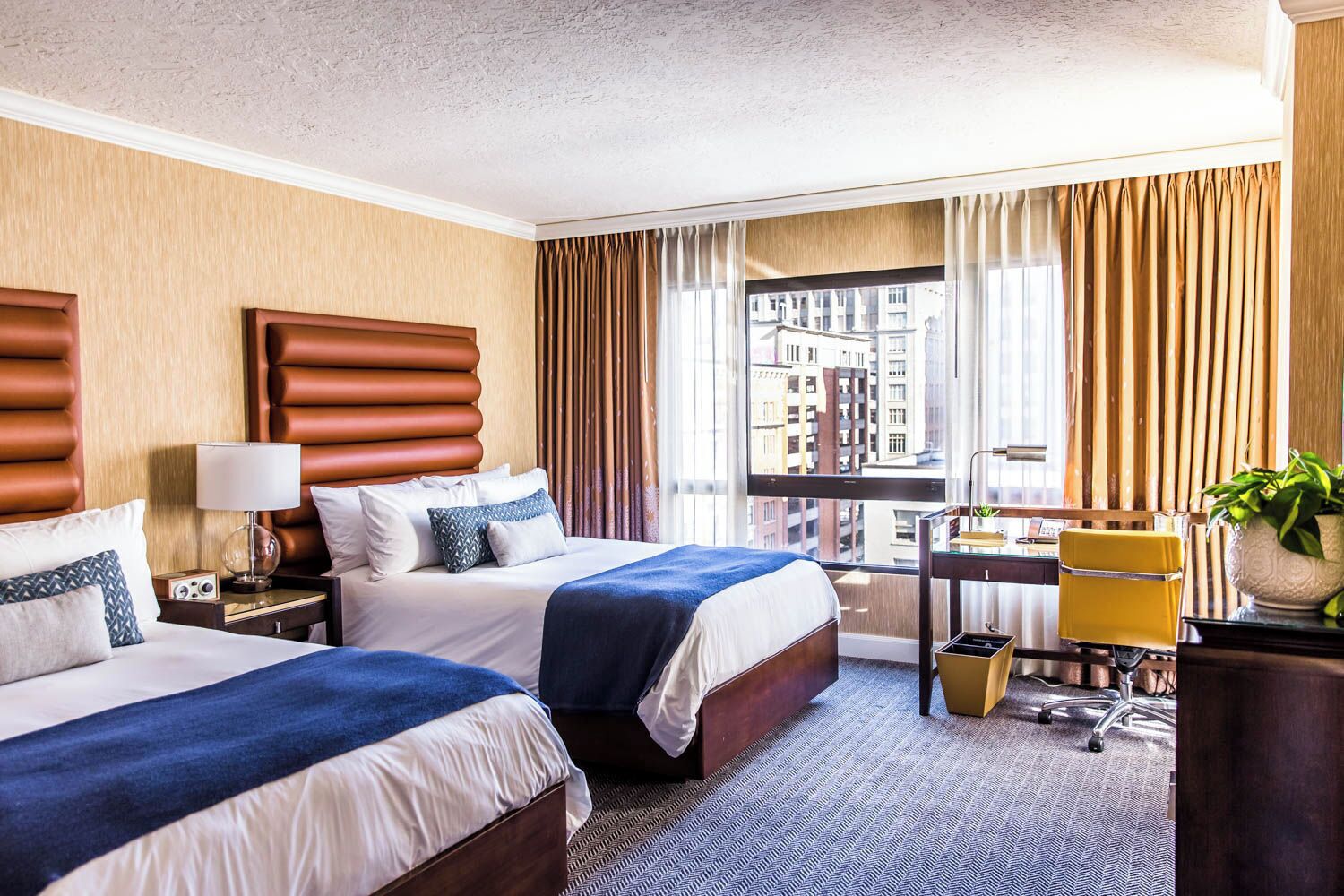 A hotel room with two beds, a desk, a chair, and a view of the city.