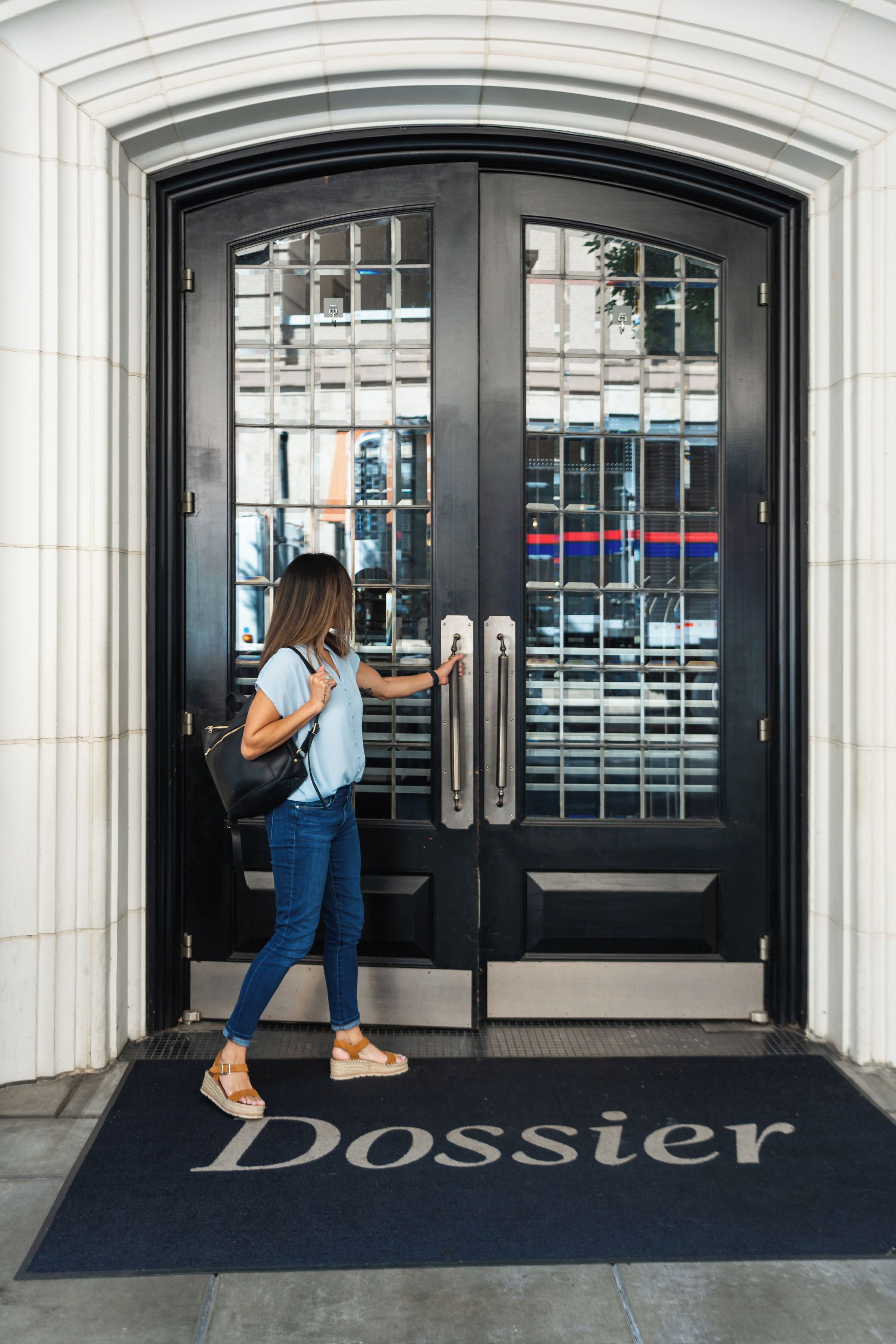 A woman approaching a black double door with a glass panel, standing on a doormat labeled "Dossier."