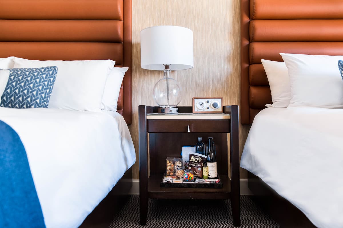 A hotel room with two beds, a nightstand with a lamp, and a tray table with snacks and drinks.