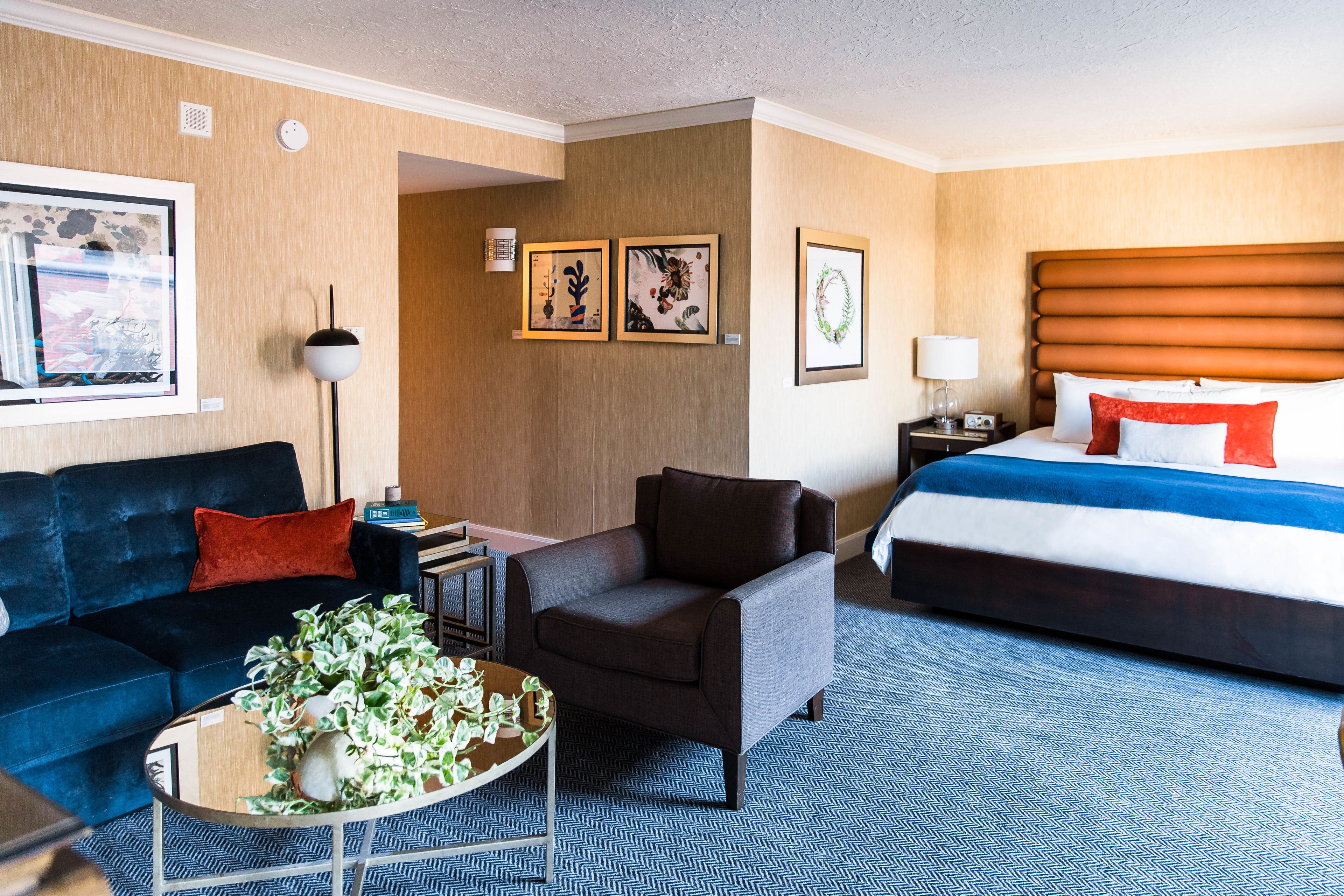 A hotel room with a bed, a sofa, a chair, a coffee table with plants, and framed artwork on the walls.