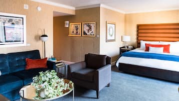 A hotel room with a bed, a sofa, a chair, a coffee table with plants, and framed artwork on the walls.