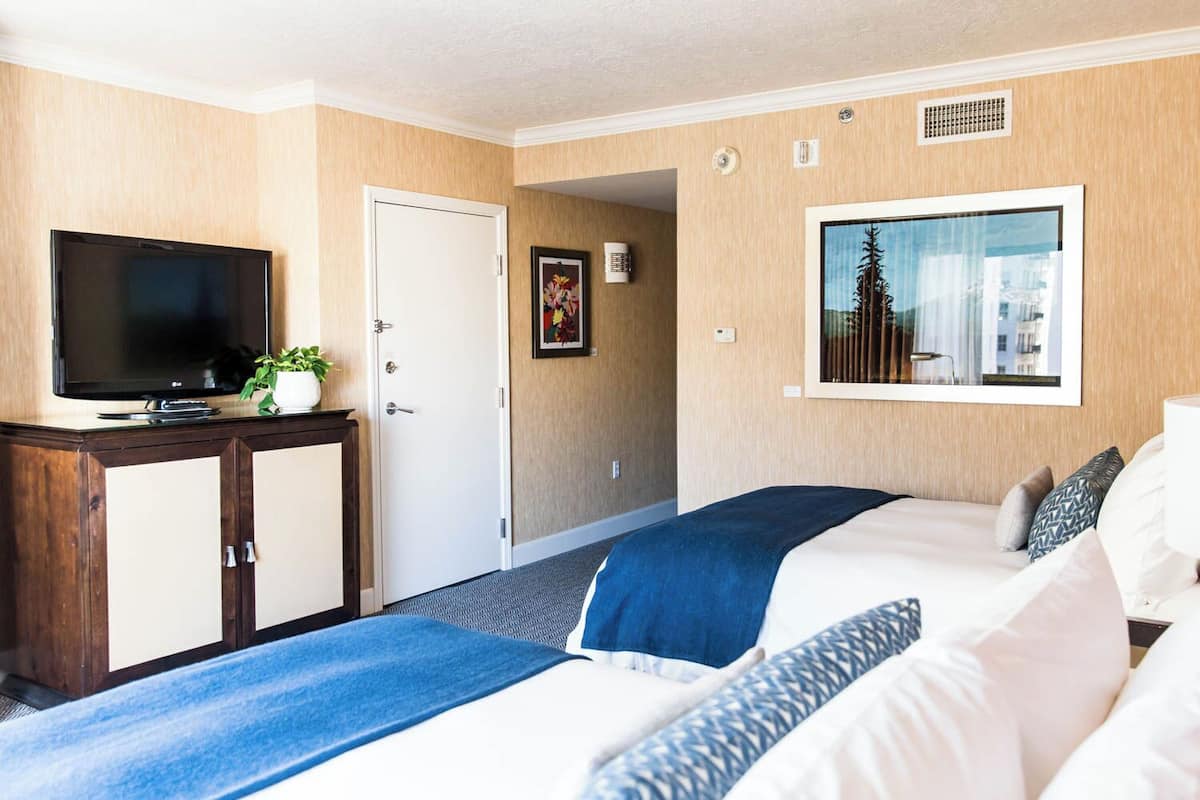 A hotel room with two beds, a television on a wooden cabinet, a framed picture on the wall, and a window with a view of trees and buildings.