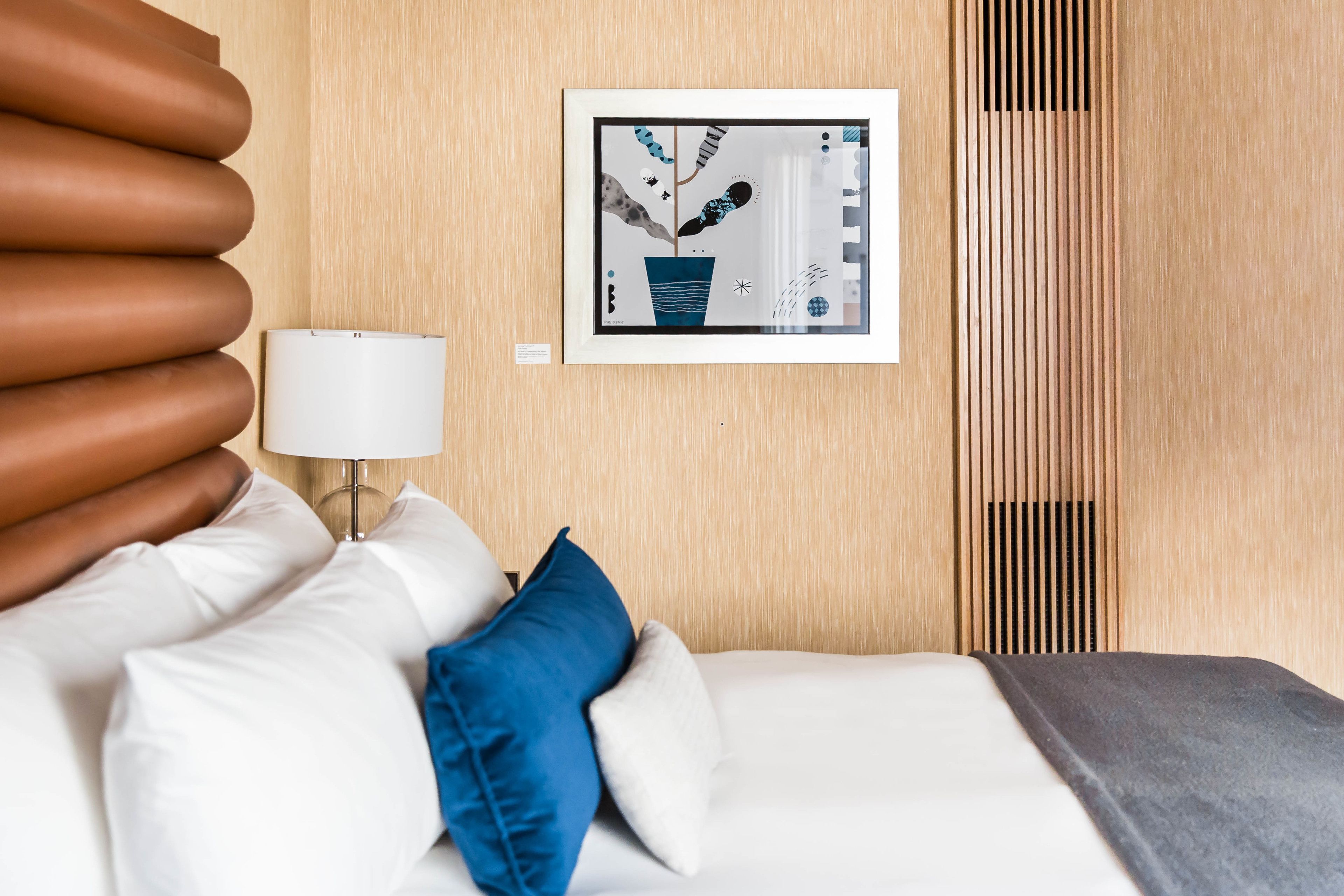 A neatly arranged hotel room with a bed, pillows, a bedside lamp, and a framed artwork on the wall.