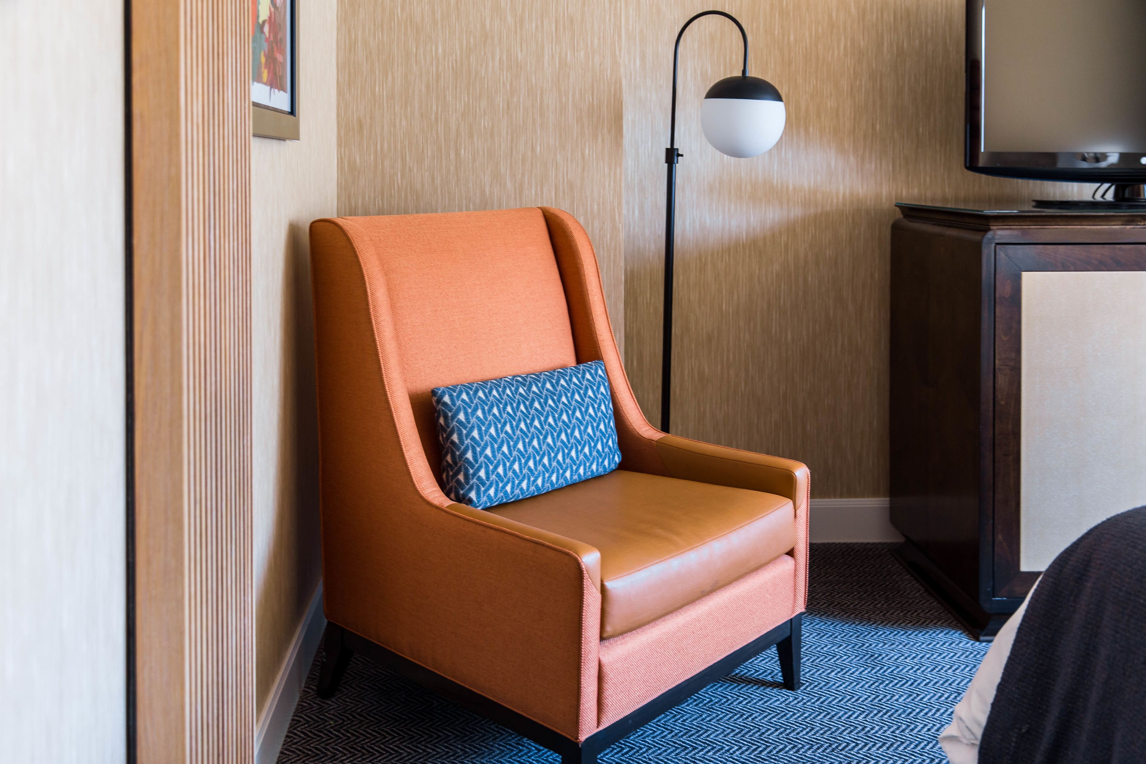 Et hotelværelse med en enkelt læderarmstol i orange, en pudde med mønster, en gulvlampe og et fjernsyn på et træskab.