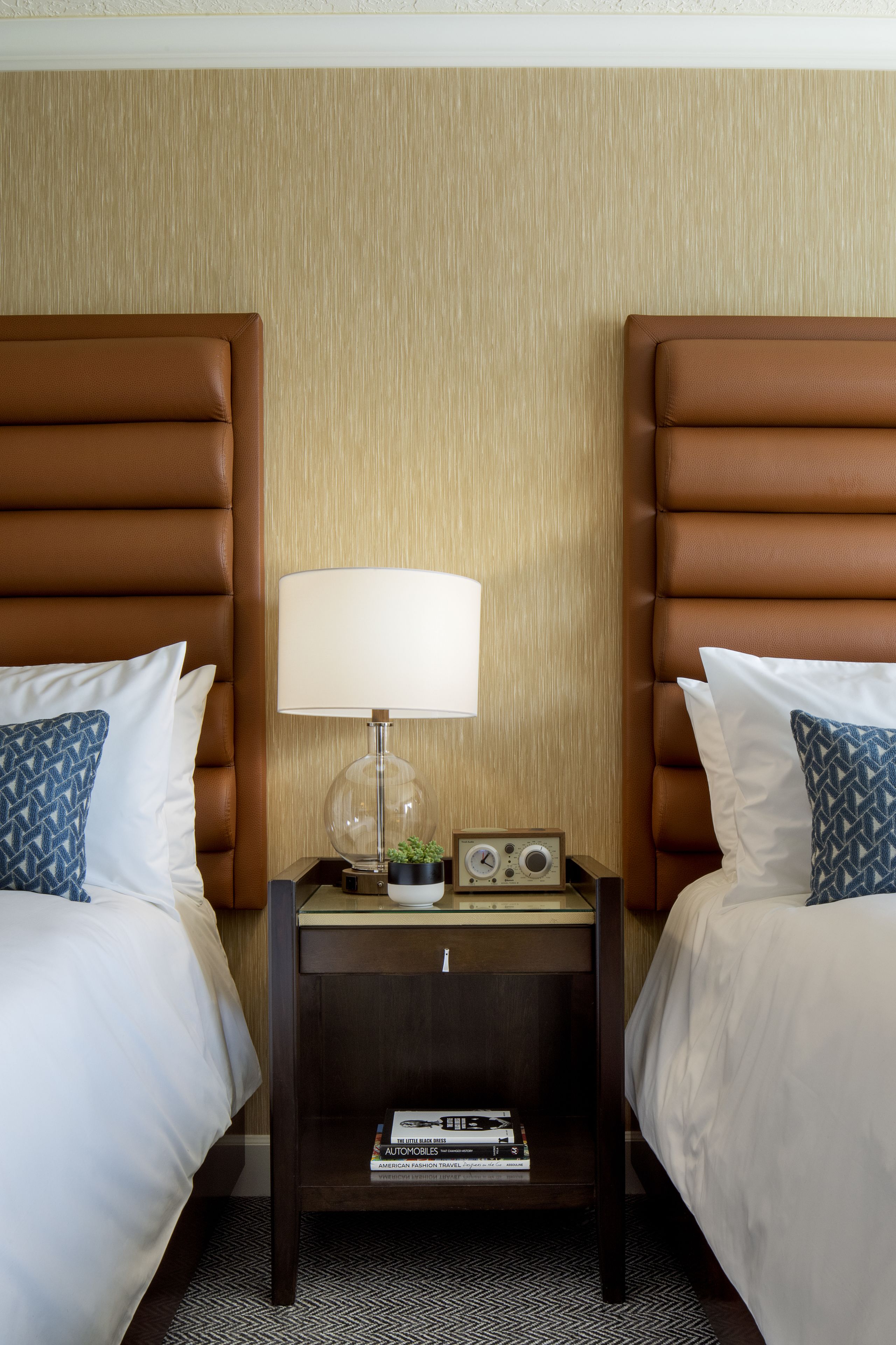 A hotel room with two beds, each with a brown headboard, white bedding, and a blue patterned pillow. There is a nightstand between the beds with a lamp, a radio, and a small plant.