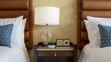 A hotel room with two beds, each with a brown headboard, white bedding, and a blue patterned pillow. There is a nightstand between the beds with a lamp, a radio, and a small plant.