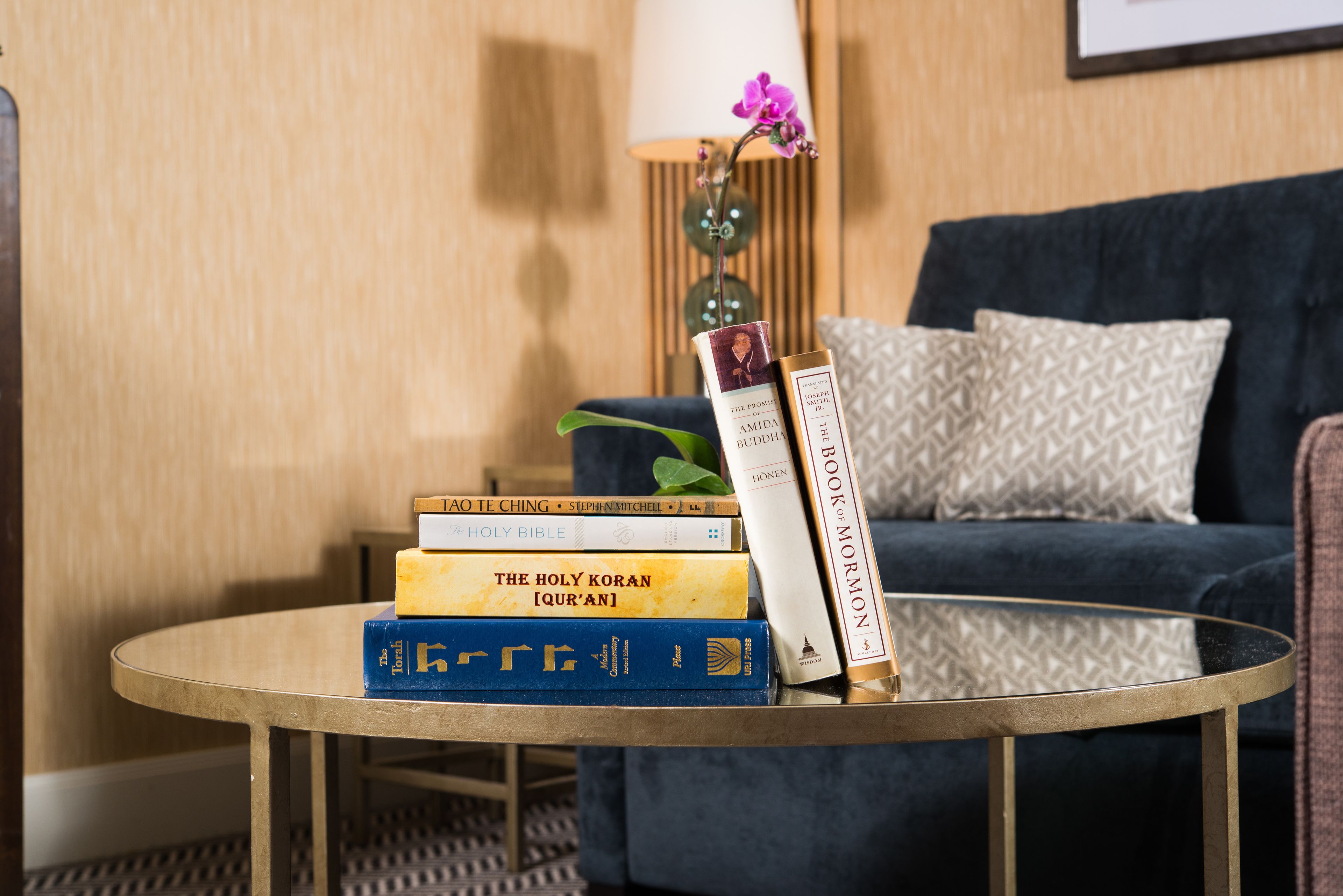A round table with a stack of books, a lamp, and a potted plant in a room with a dark sofa and patterned cushions.