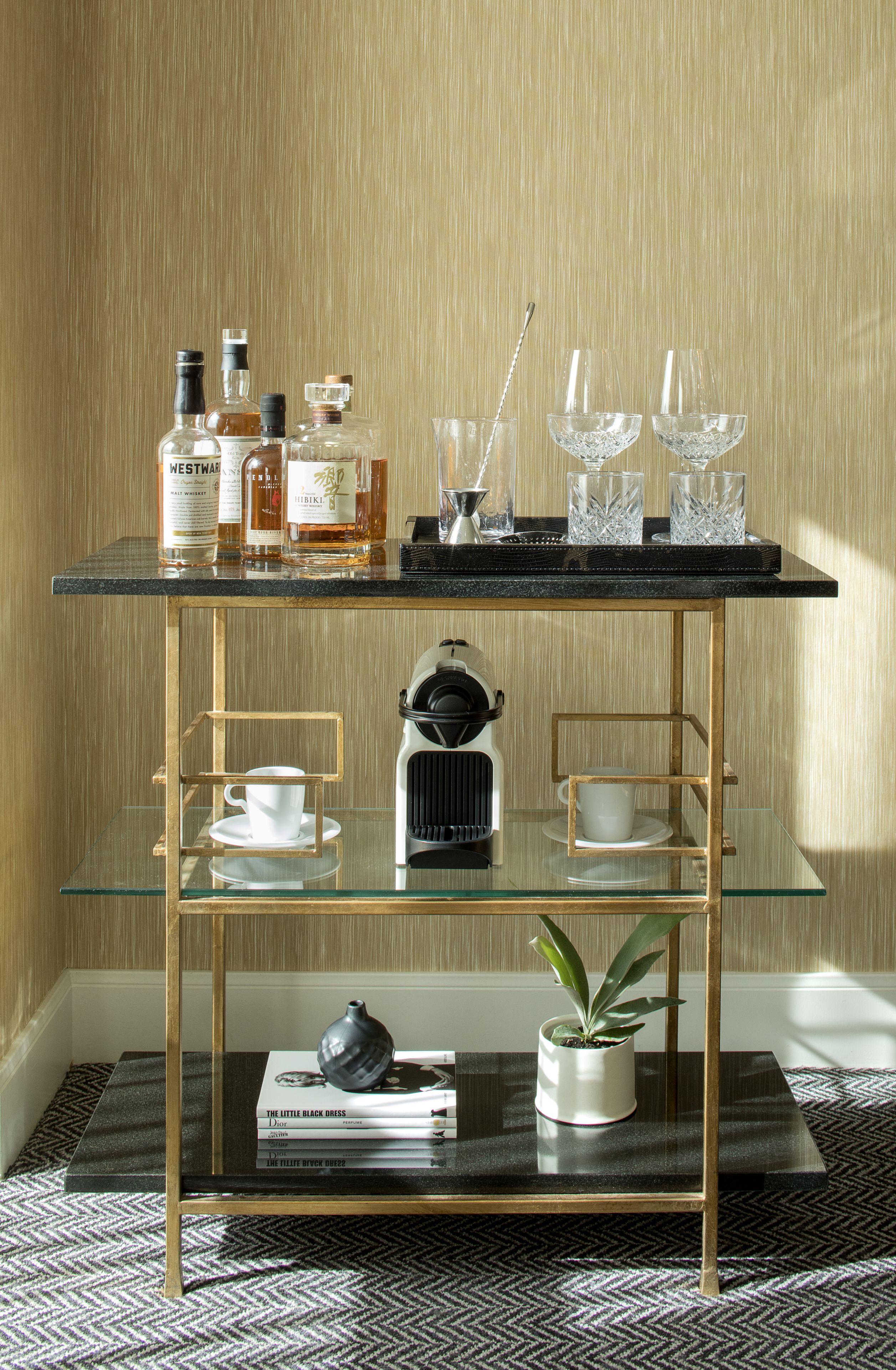 A glass coffee table with a black top and gold frame, holding a coffee machine, cups, and a potted plant.