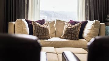 Ein Wohnzimmer mit einem beigefarbenen Sofa, gemusterten Kissen und Blick durch ein mit Vorhängen drapiertes Fenster.