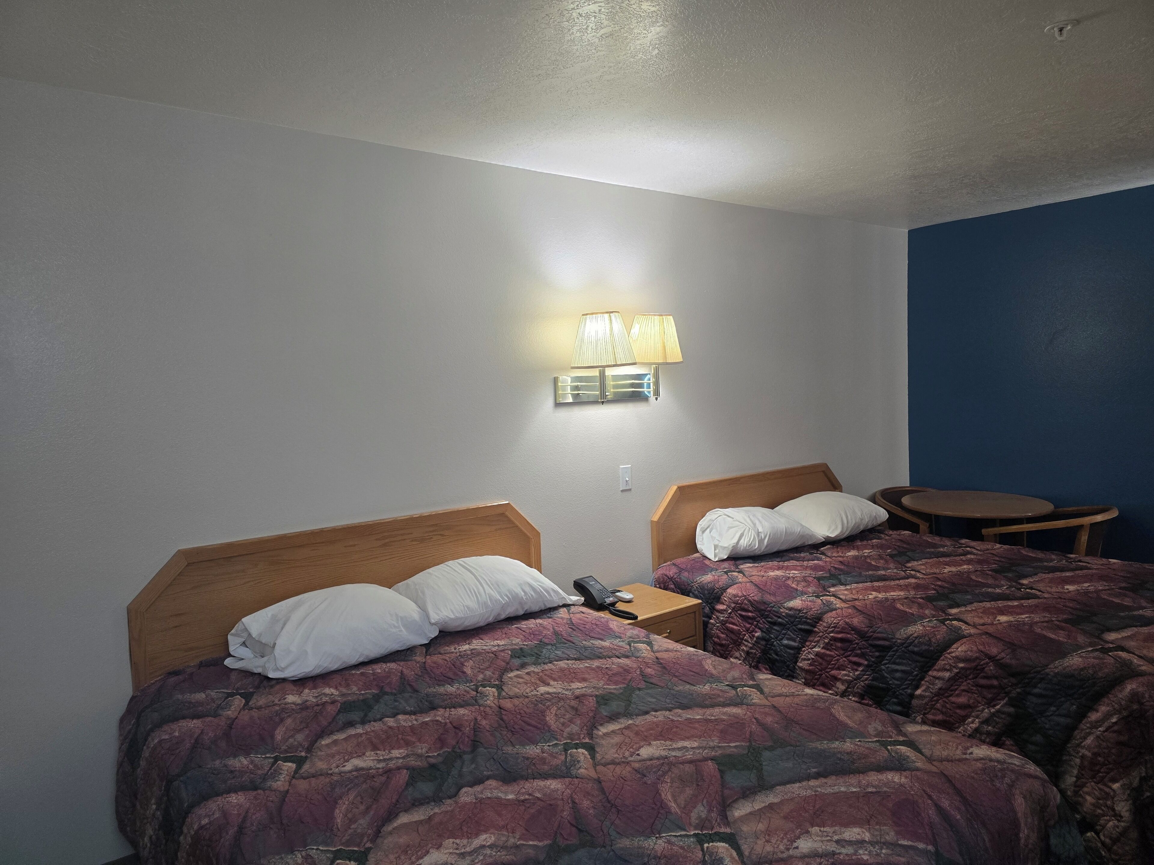 A hotel room with two beds, a bedside table with a phone, a lamp, and a small round table.