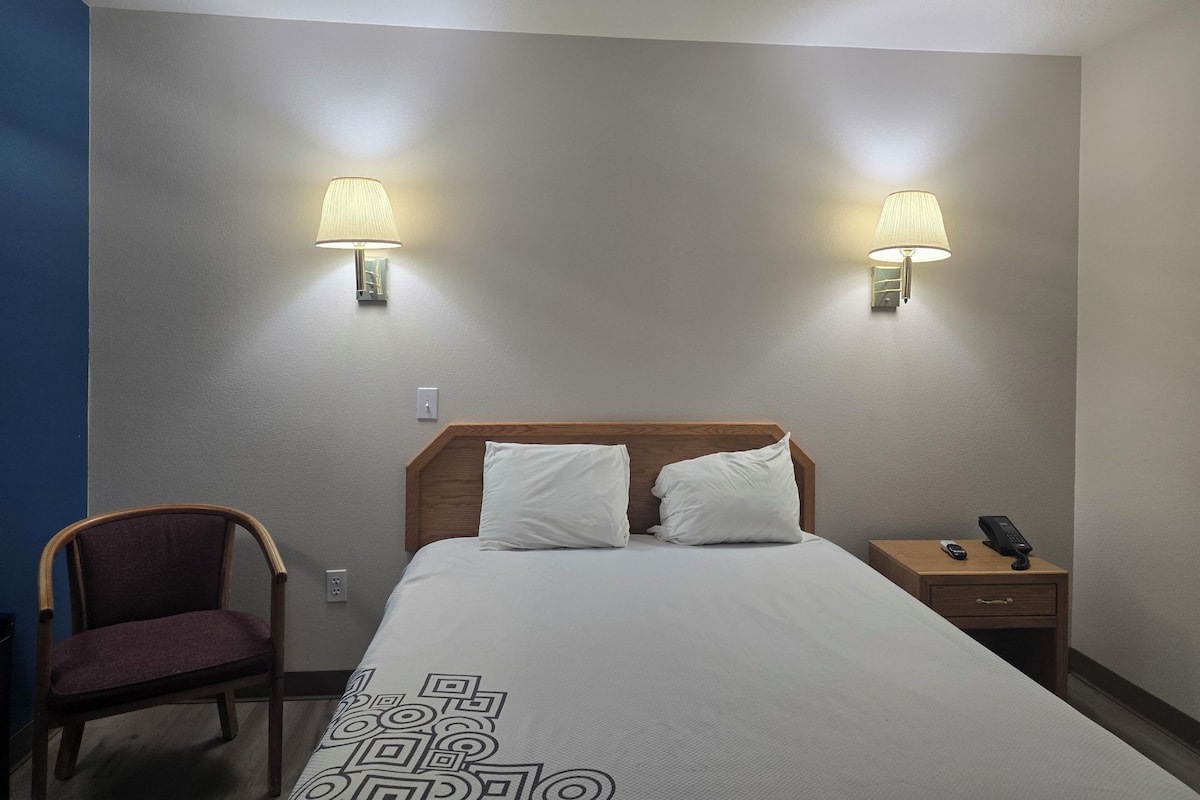 A hotel room with a bed, two bedside lamps, a chair, a nightstand with a phone, and a wall socket.