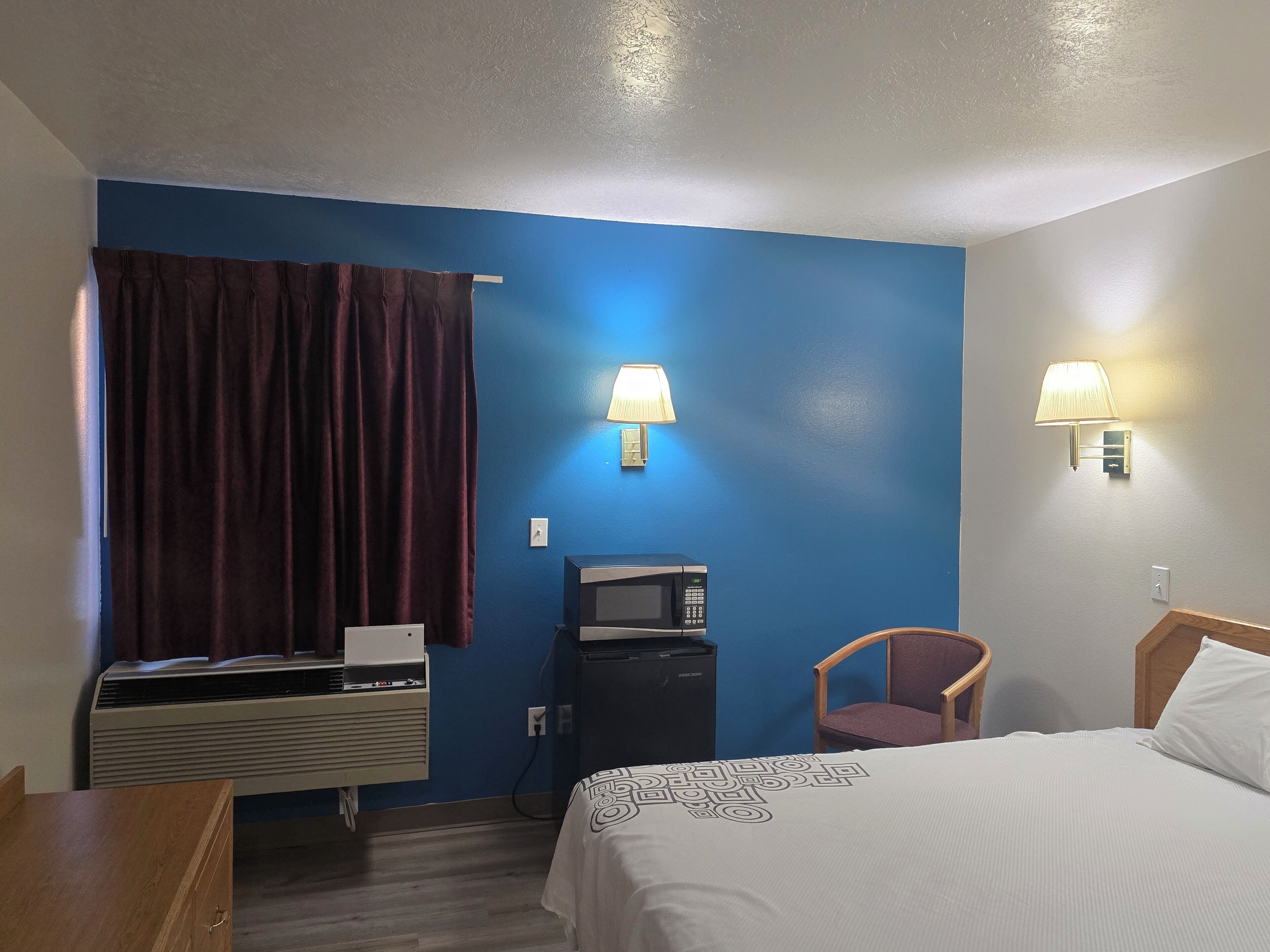 Een hotelkamer met een bed, een bureau, een stoel, een magnetron, een airconditioner en twee wandlampen.