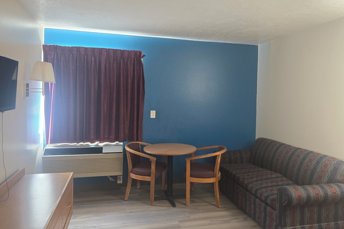 A room with a blue wall, a sofa, a table with two chairs, a lamp, and a window with curtains.