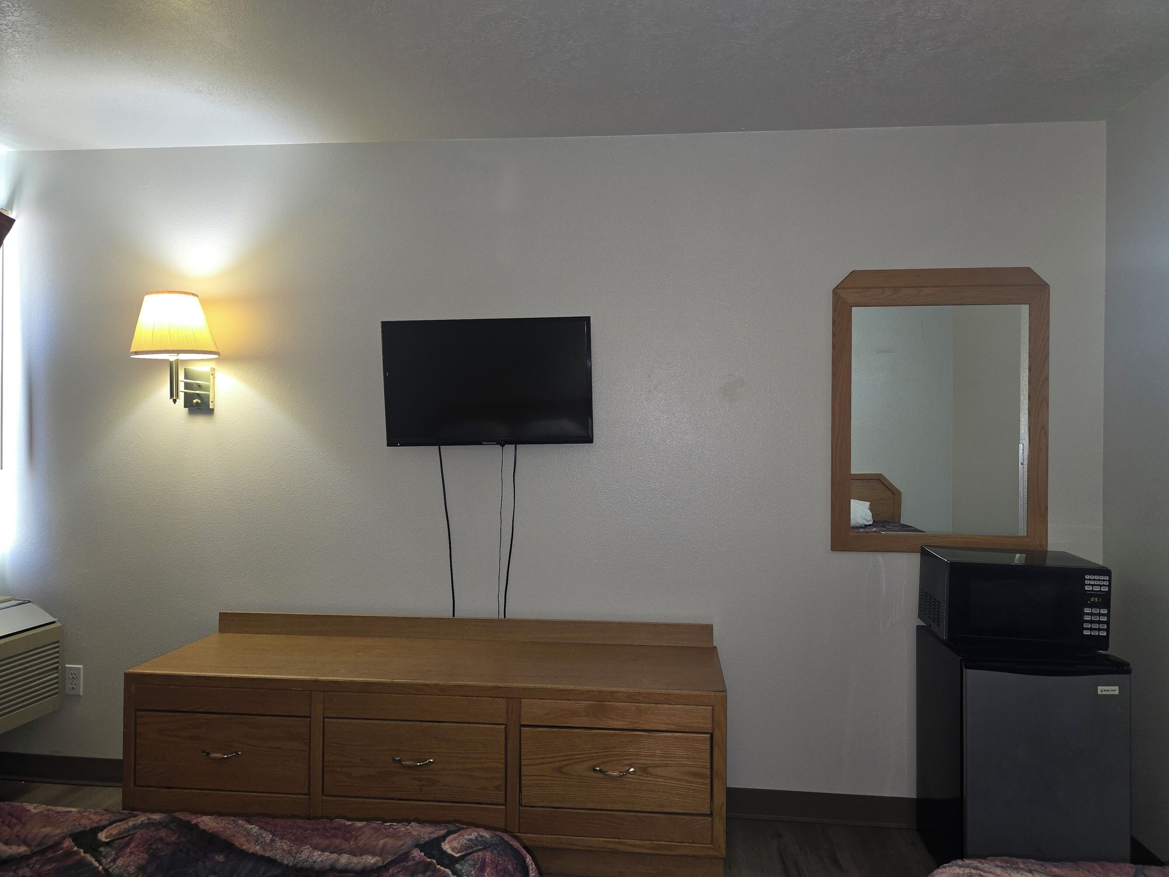 A hotel room with a flat-screen TV mounted on the wall, a wooden dresser, a microwave, and a wall-mounted lamp.