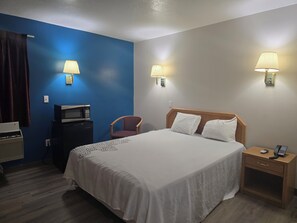 A hotel room with a bed, two bedside lamps, a nightstand with a phone, a microwave, a chair, and a curtain.