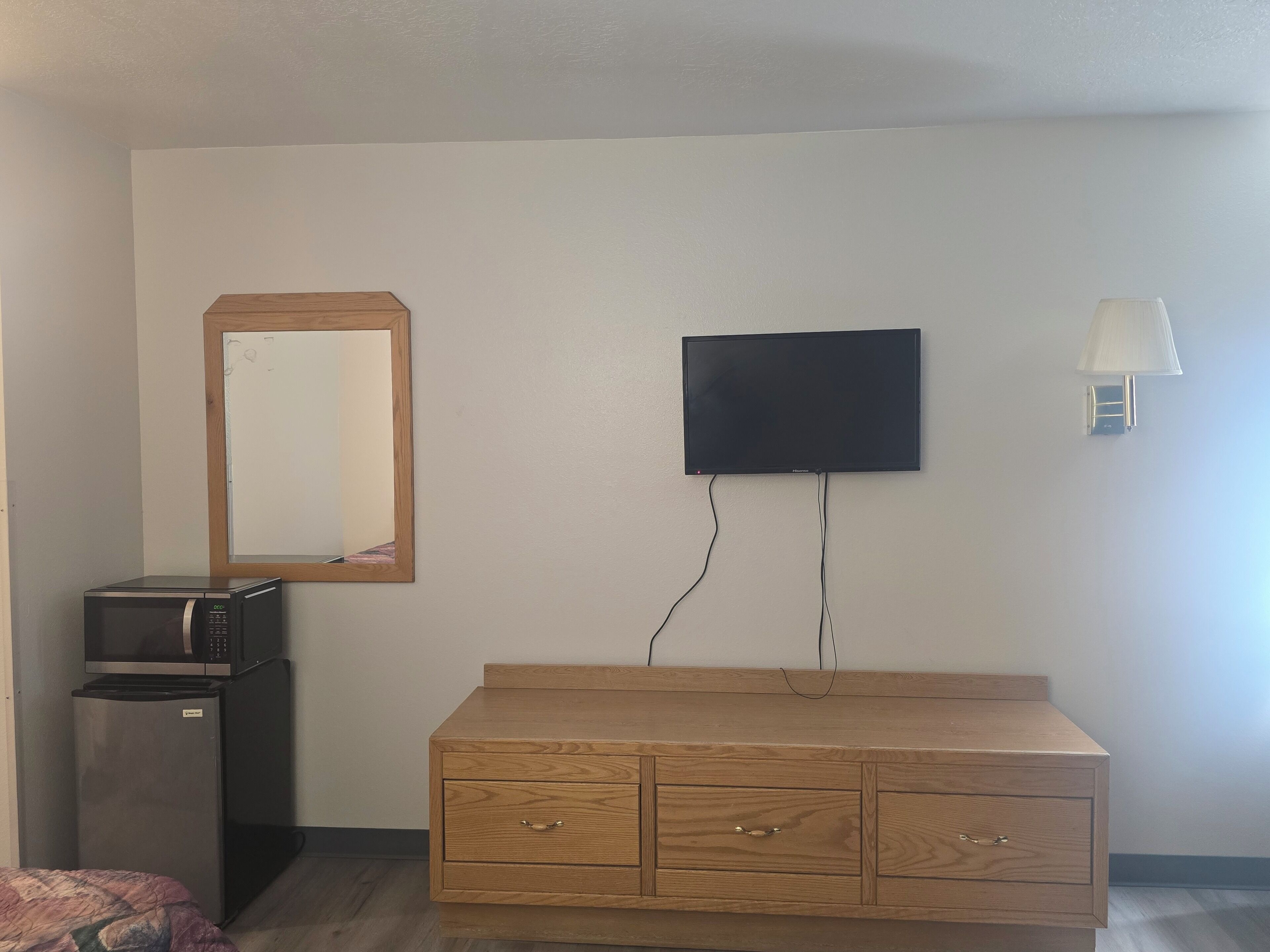 A room with a wooden dresser, a flat-screen TV mounted on the wall, a microwave, and a small refrigerator.