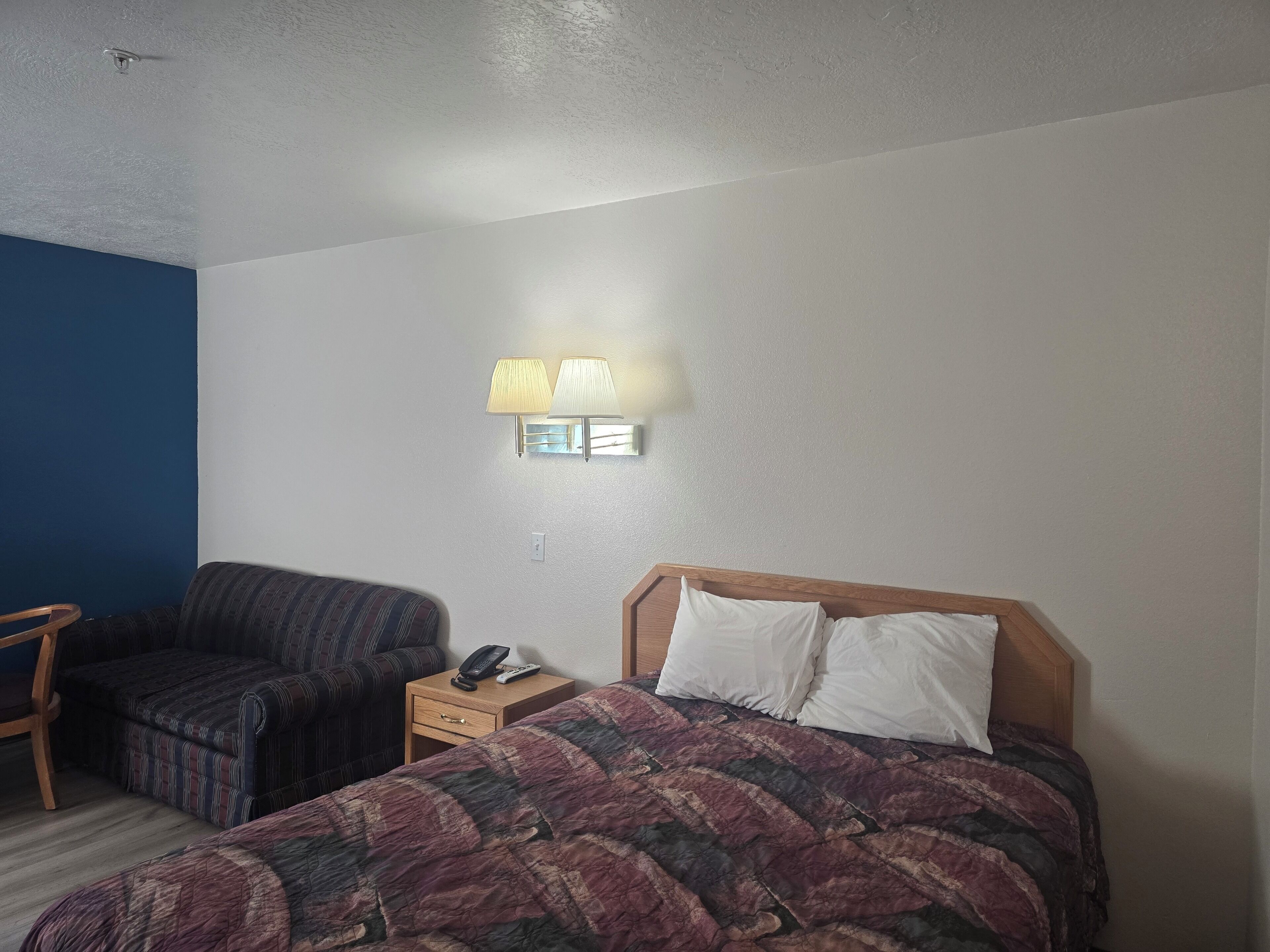 A hotel room with a bed, a chair, a nightstand, and a wall-mounted lamp.