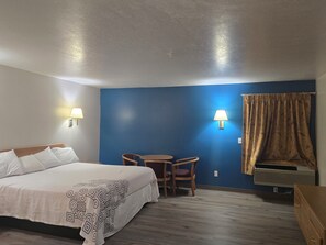 A hotel room with a bed, a small table with chairs, a wall-mounted lamp, and a window with curtains.