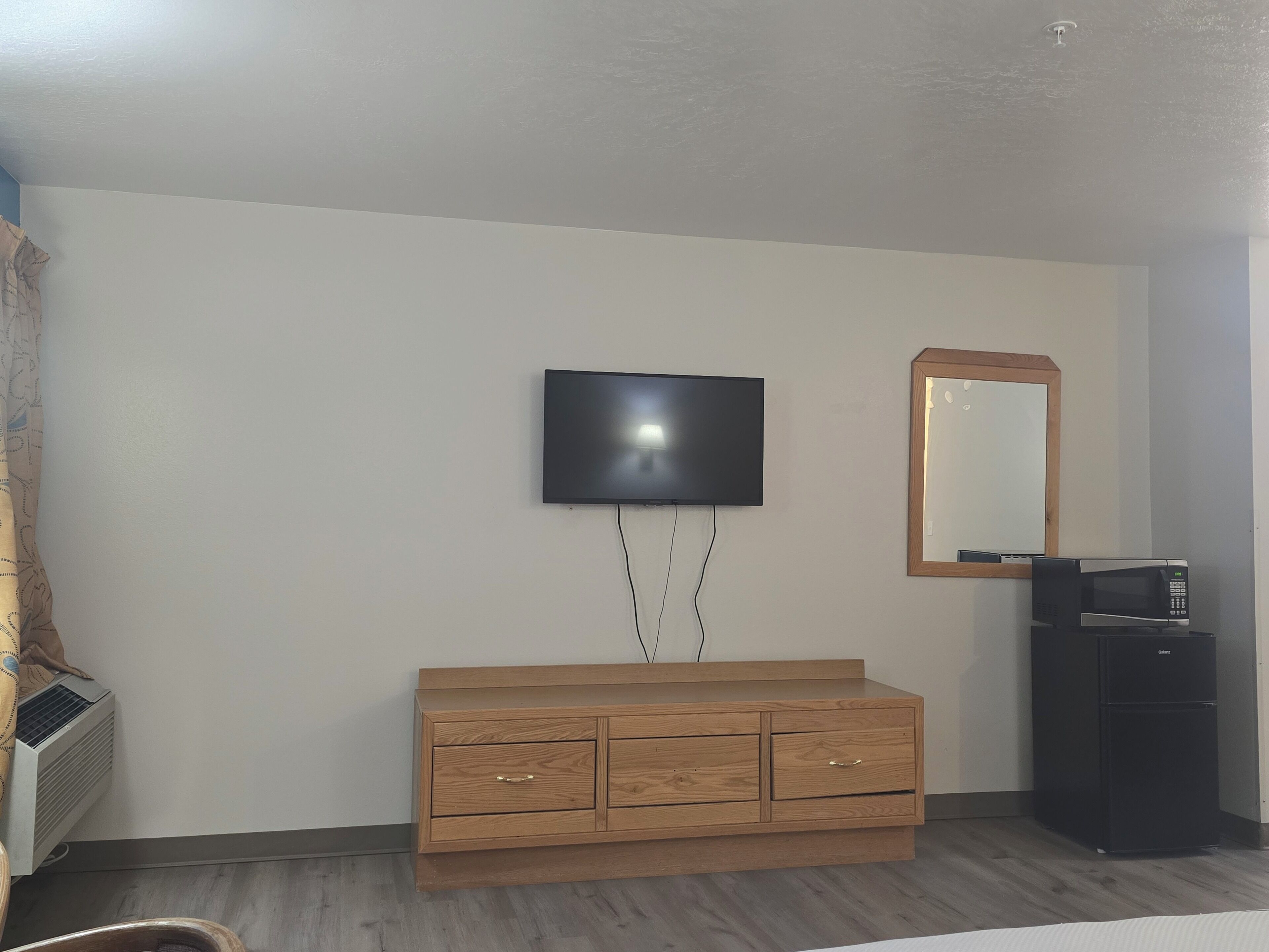 A room with a flat-screen TV mounted on the wall, a wooden dresser with drawers, a mirror with a wooden frame, a black refrigerator, and an air conditioning unit.