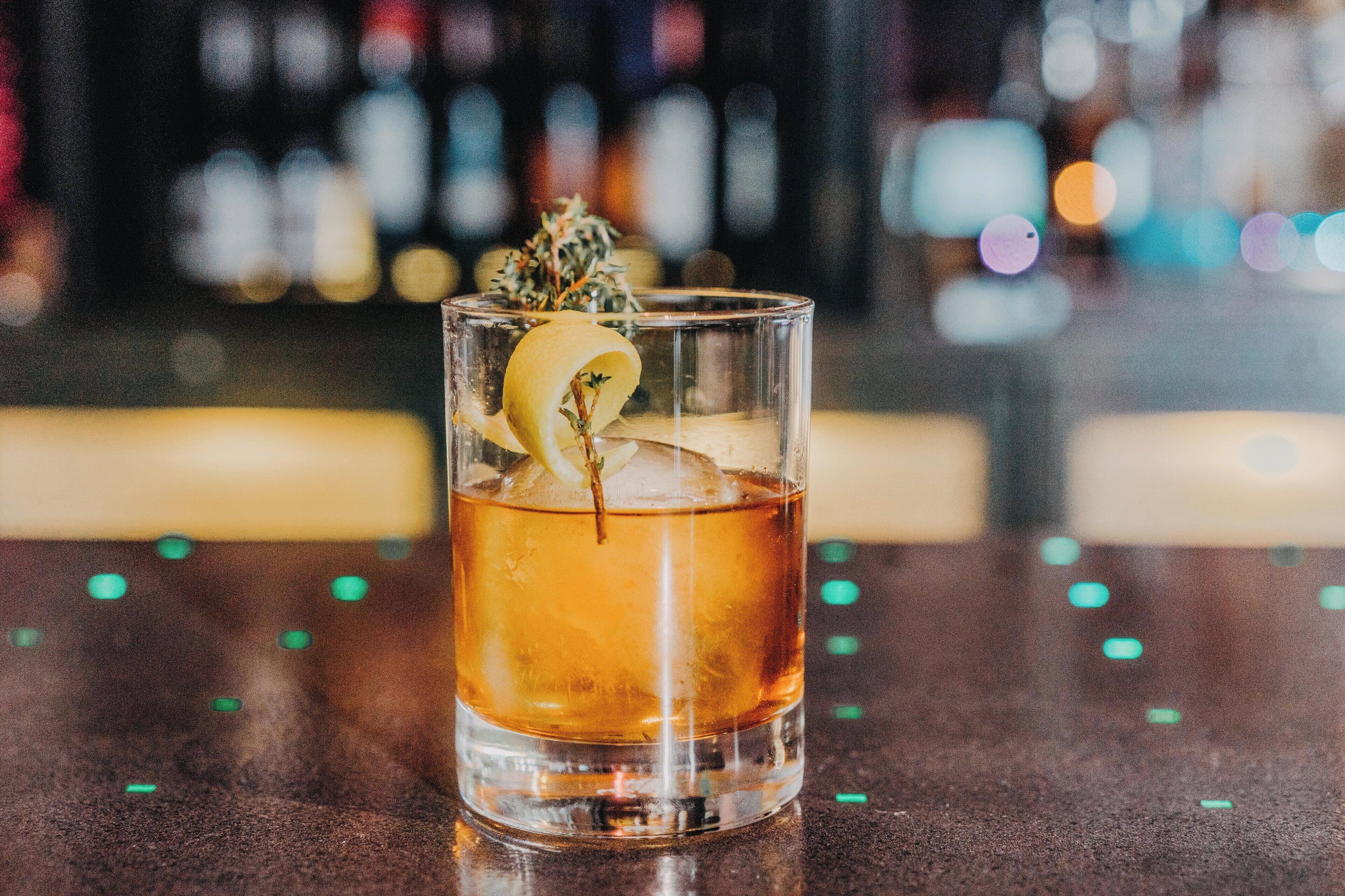 A glass of amber-colored beverage with a lemon slice and a sprig of herb, placed on a reflective surface with blurred background featuring bottles.