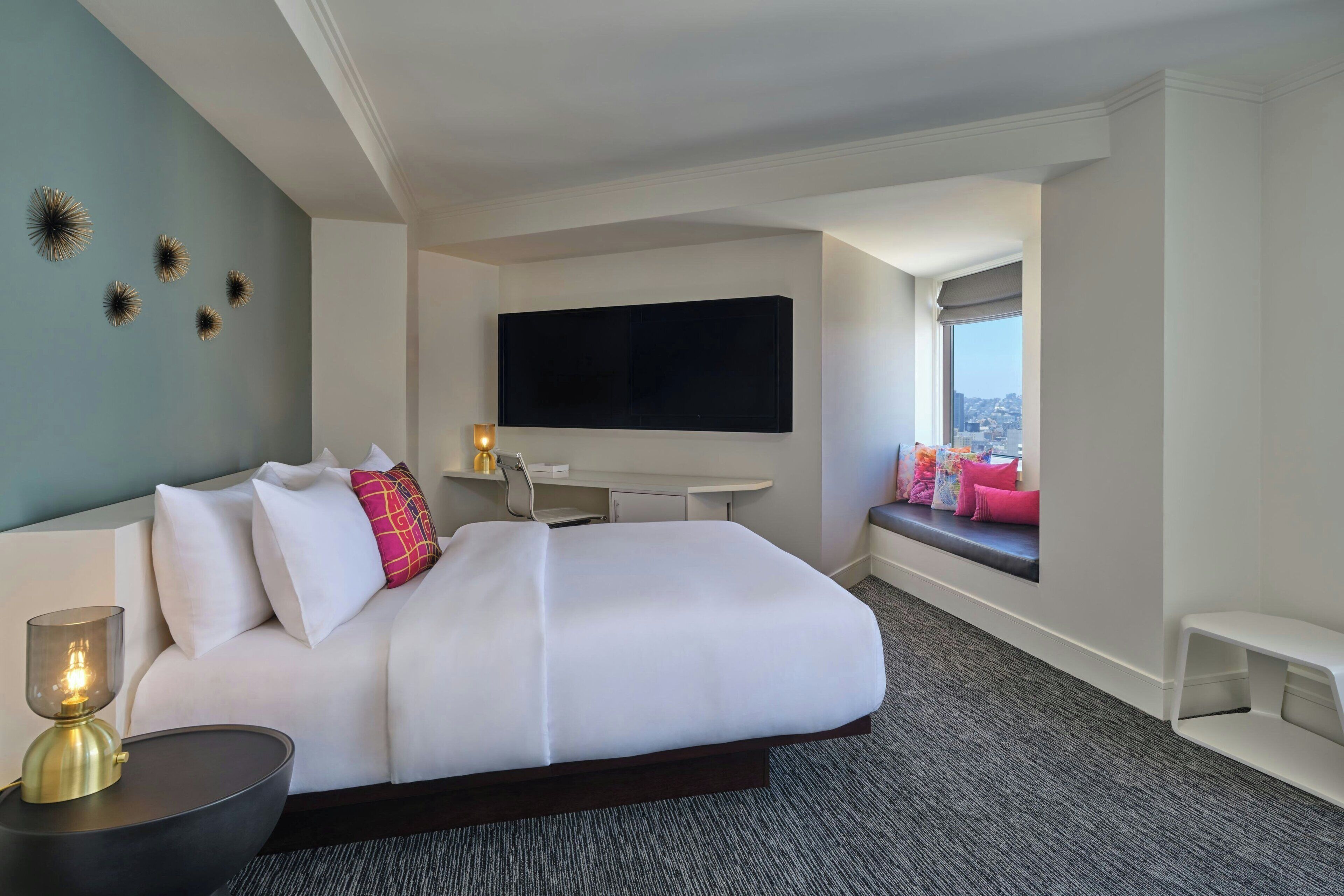 Quarto de hotel moderno com uma cama grande, uma TV de tela plana na parede, uma escrivaninha e uma janela com vista para a cidade.