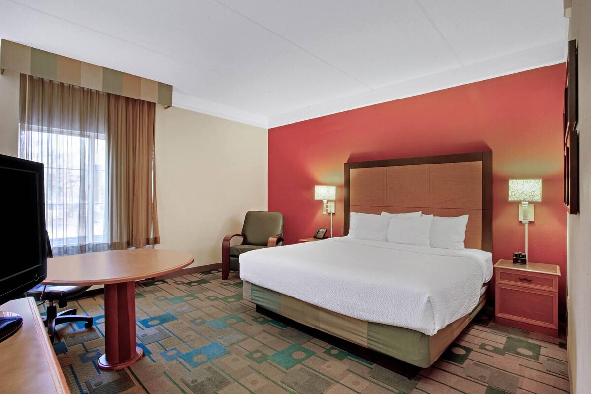 A hotel room with a large bed, a desk, a chair, a TV, and a nightstand with a lamp.
