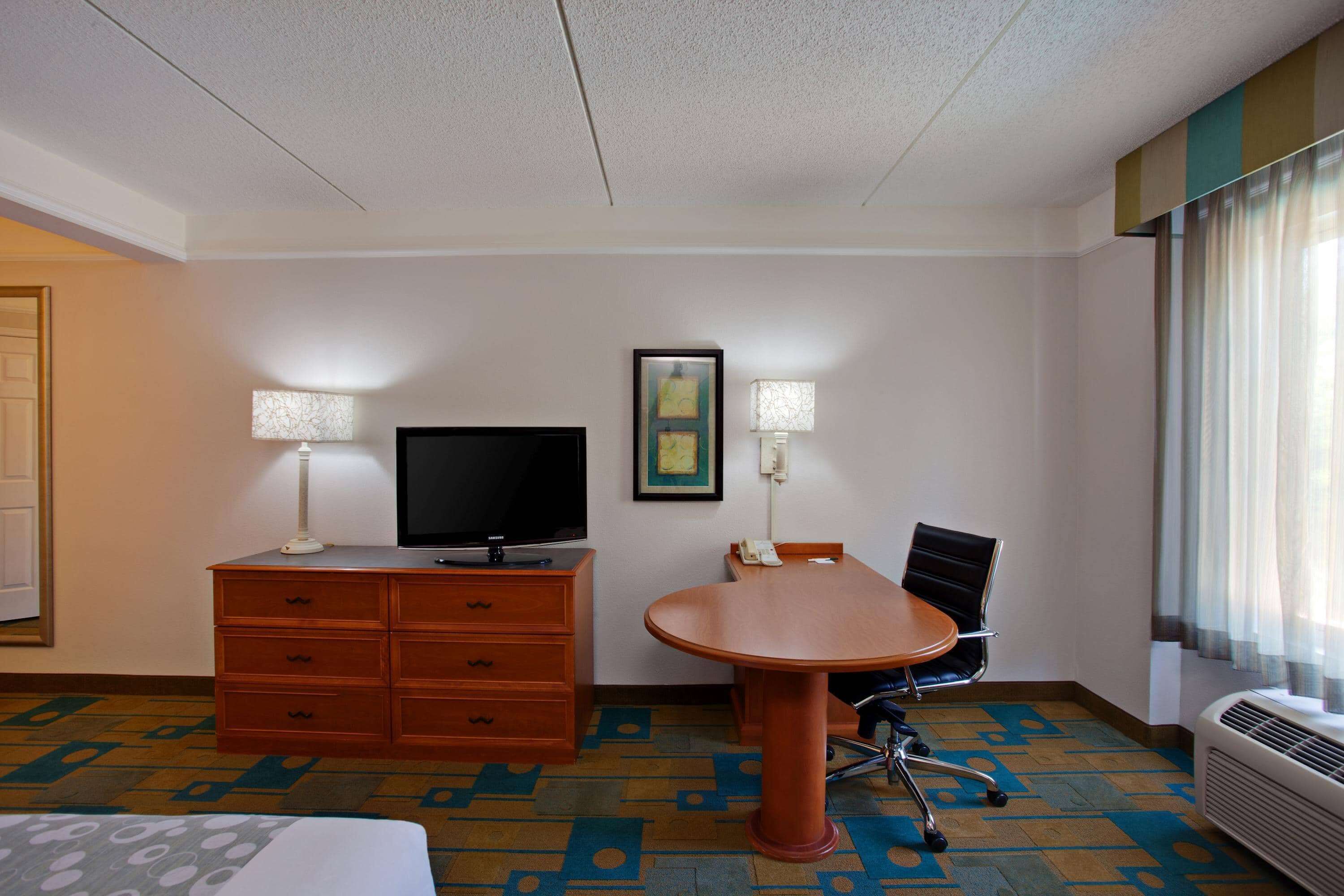 A hotel room with a bed, a desk with a chair, a television, and a desk lamp.