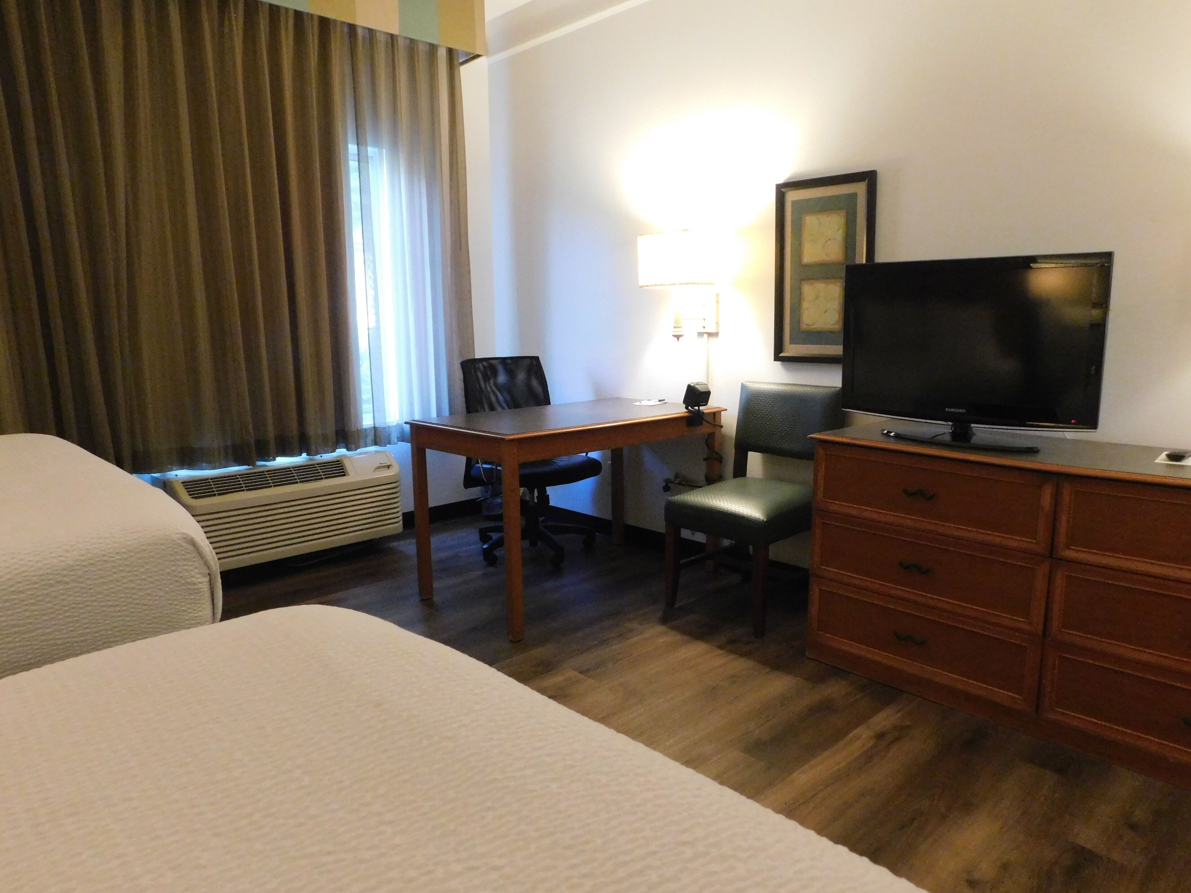 Une chambre d’hôtel avec deux lits, un bureau avec une chaise, une télévision, une commode et une fenêtre avec des rideaux.