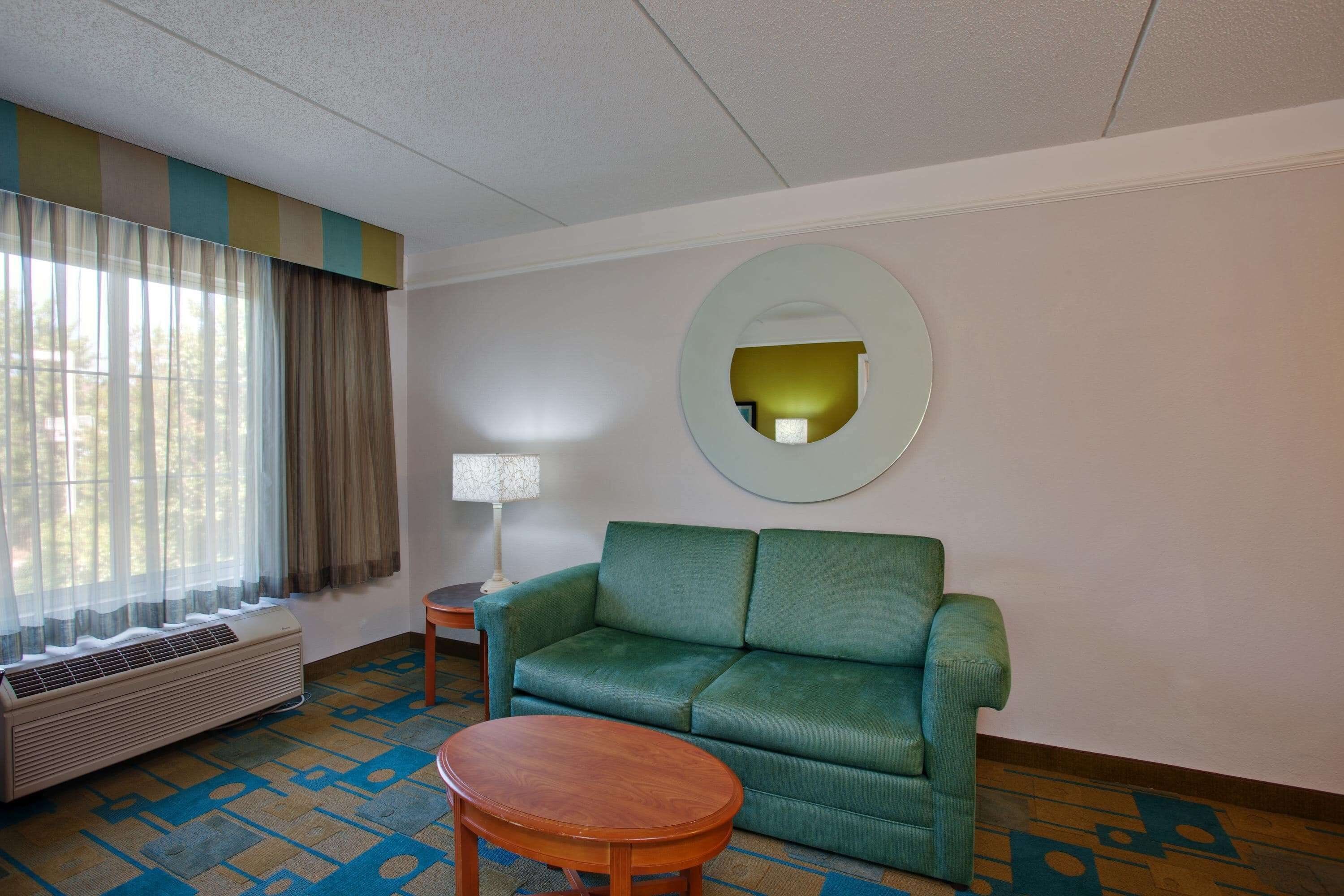 A hotel room with a green sofa, a wooden coffee table, a round mirror, and a window with curtains.