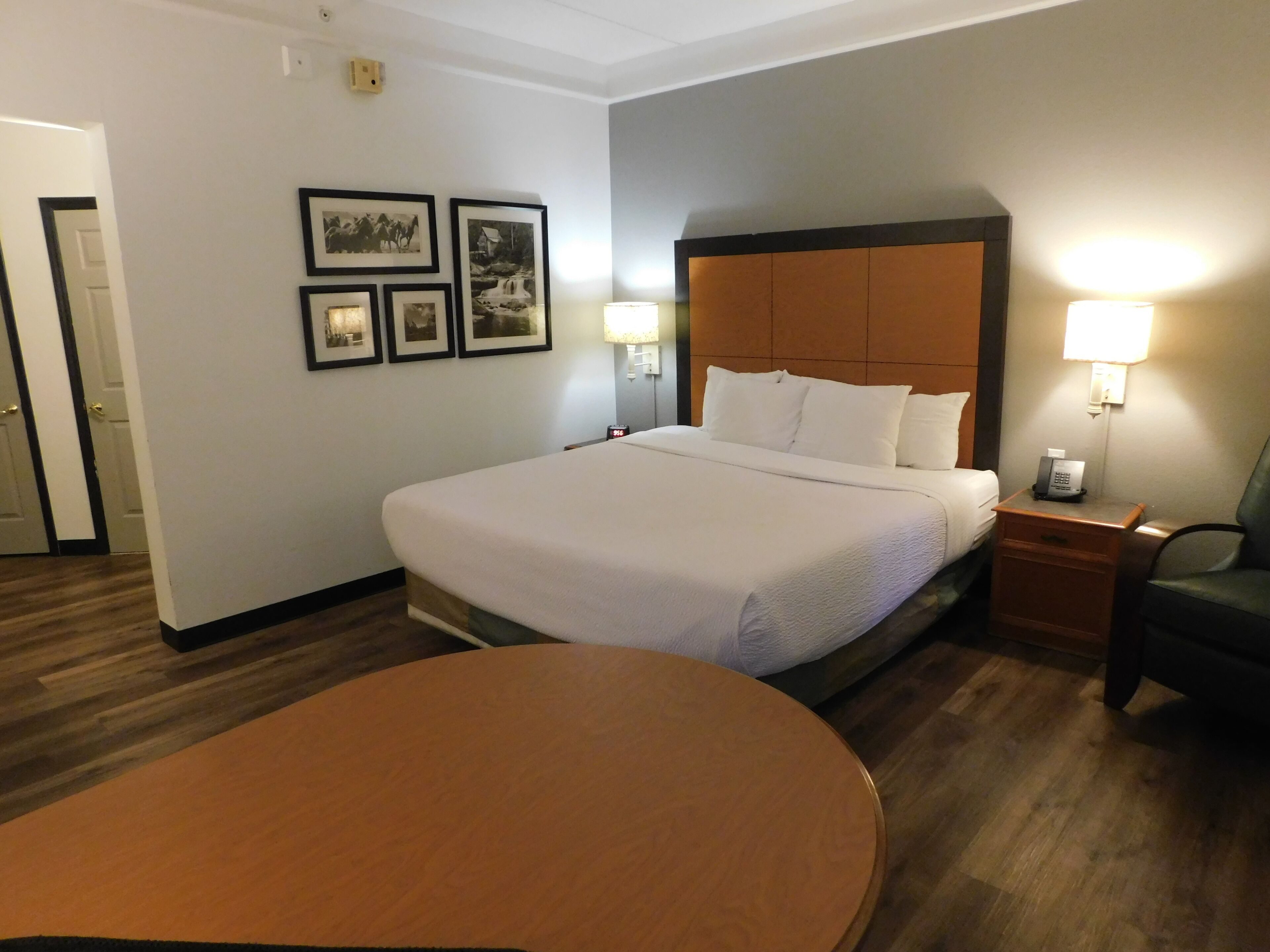 Une chambre d’hôtel avec un grand lit, une table de chevet avec une lampe, une chaise et une table ronde. Des tableaux encadrés ornent le mur.