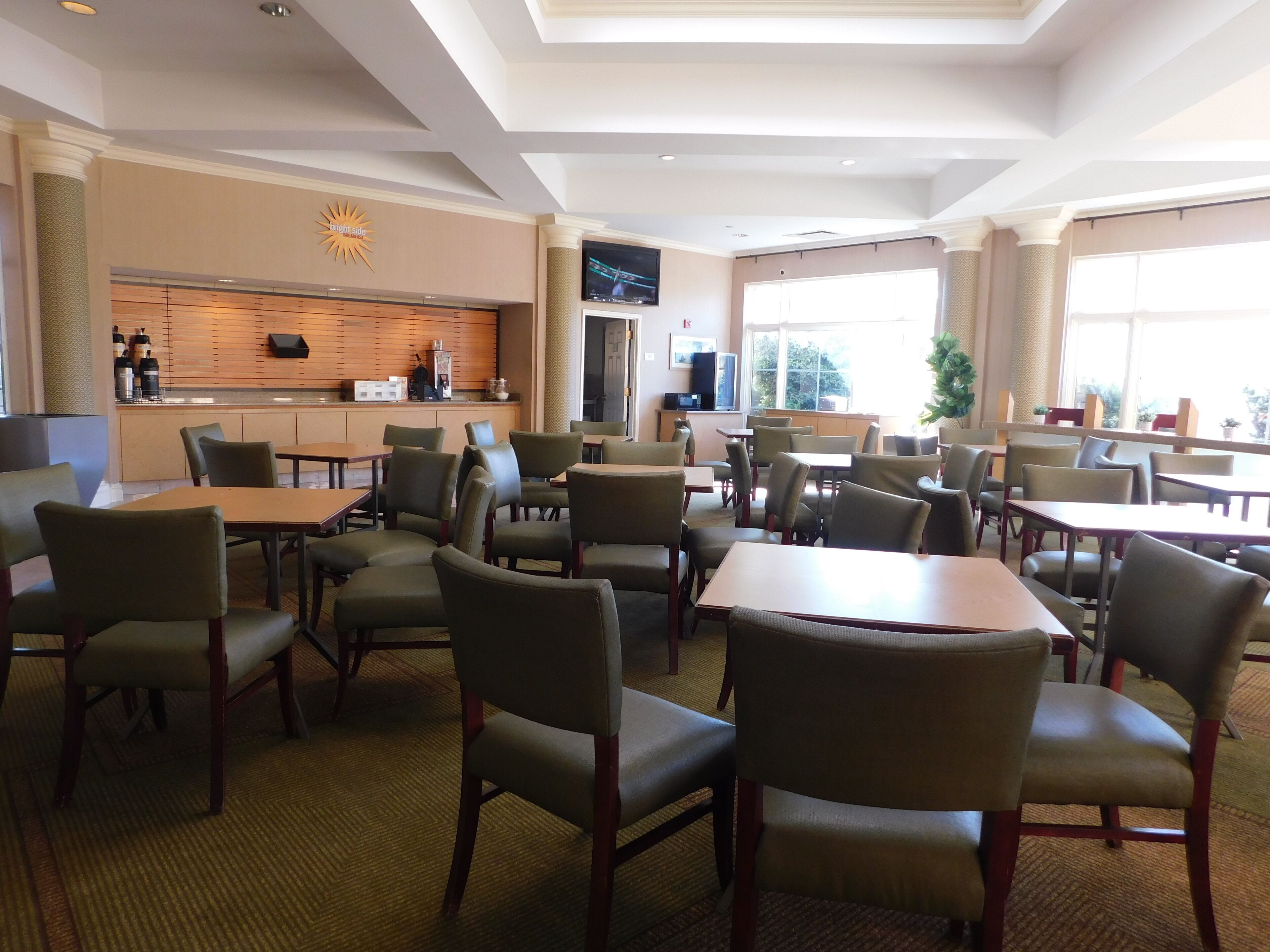 A hotel dining area with multiple tables and chairs, a bar area with a TV, and large windows providing natural light.