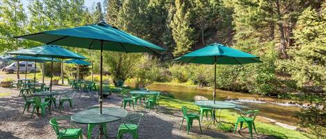 An outdoor seating area with green tables and chairs, surrounded by trees and a river.
