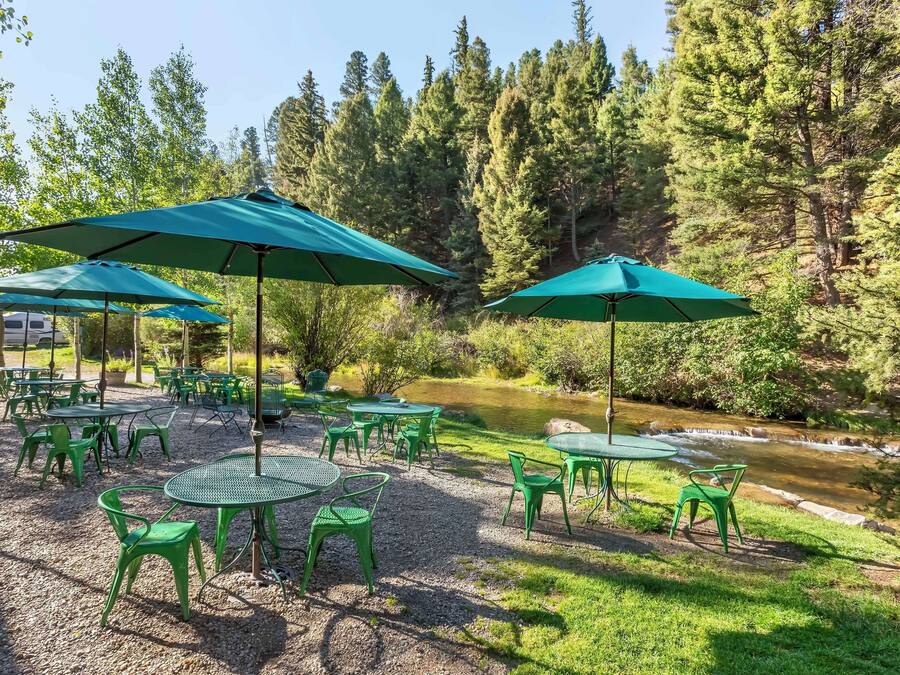 An outdoor seating area with green tables and chairs, surrounded by trees and a river.