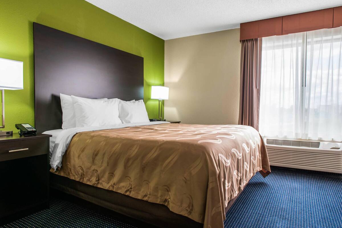Une chambre d’hôtel avec un grand lit, deux tables de chevet avec des lampes, une fenêtre avec des rideaux et un appareil de climatisation.