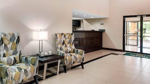 Un hall d’hôtel avec deux fauteuils à motifs, une petite table avec une lampe, un comptoir d’accueil et une grande porte vitrée donnant sur l’extérieur.