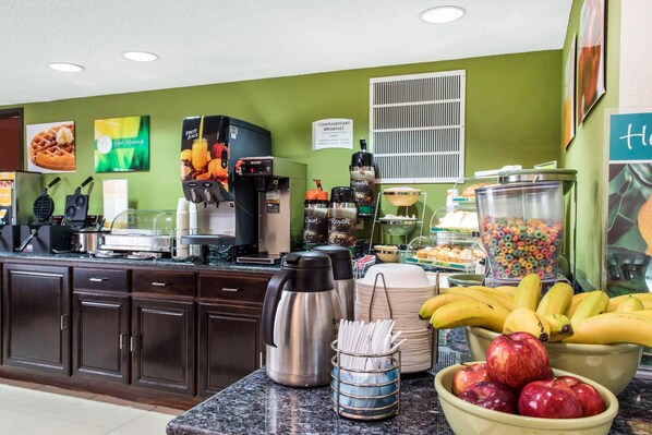 Una barra de desayuno con máquinas de café, un dispensador de fruta y una variedad de frutas y cereales.