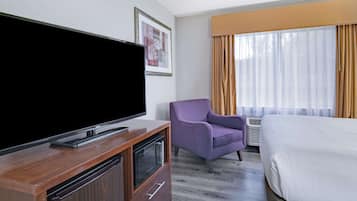 A hotel room with a flat-screen TV on a wooden stand, a purple armchair, a bed with white bedding, and a large window with curtains.