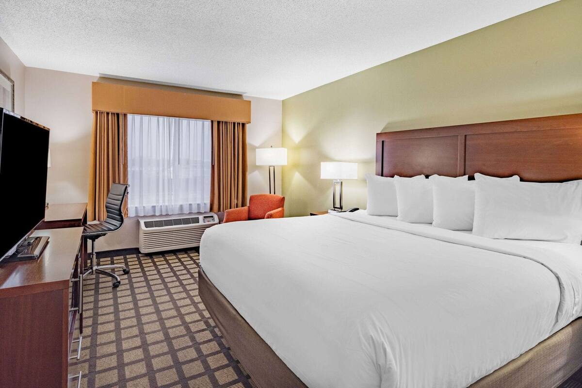 A hotel room with a large bed, a desk with a chair, an air conditioning unit, a television, and a window with curtains.