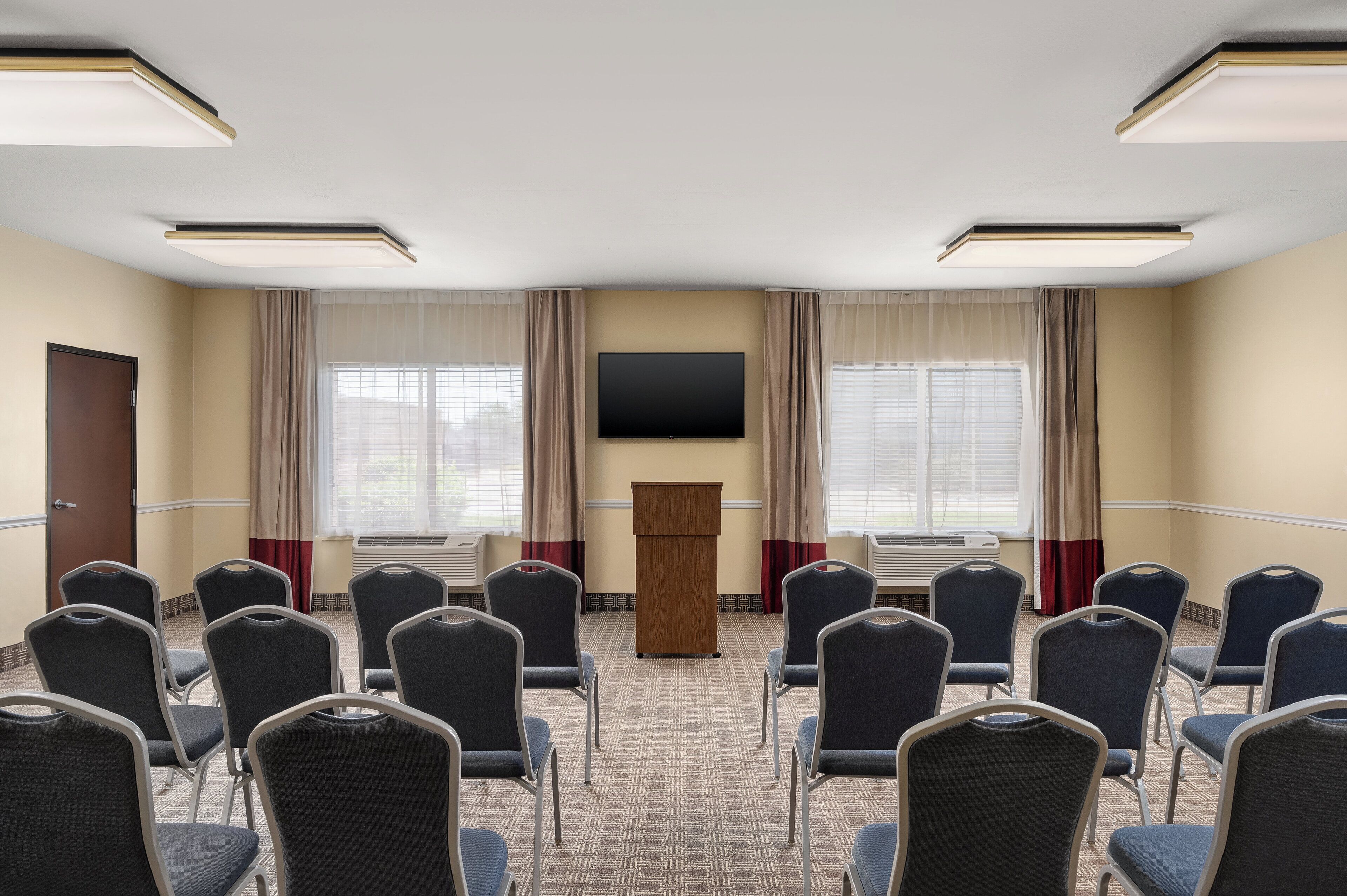 A conference room with rows of chairs, a podium, a flat-screen TV, and large windows with curtains.