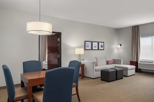 Una habitación de hotel con una mesa de comedor, sillas, un sofá, una lámpara y una ventana con cortinas.