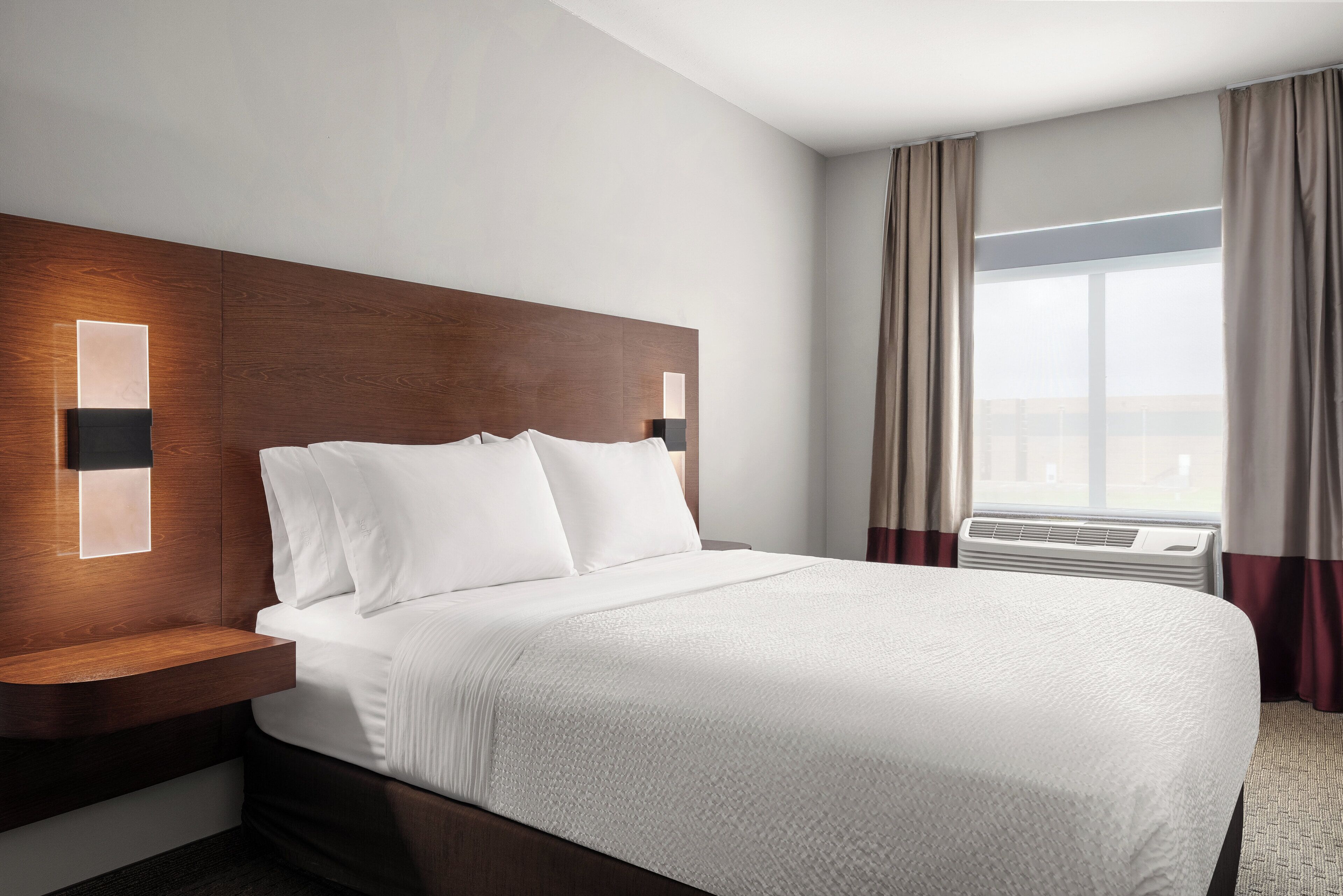 A hotel room with a large bed, a wooden headboard, a nightstand, and a window with curtains.
