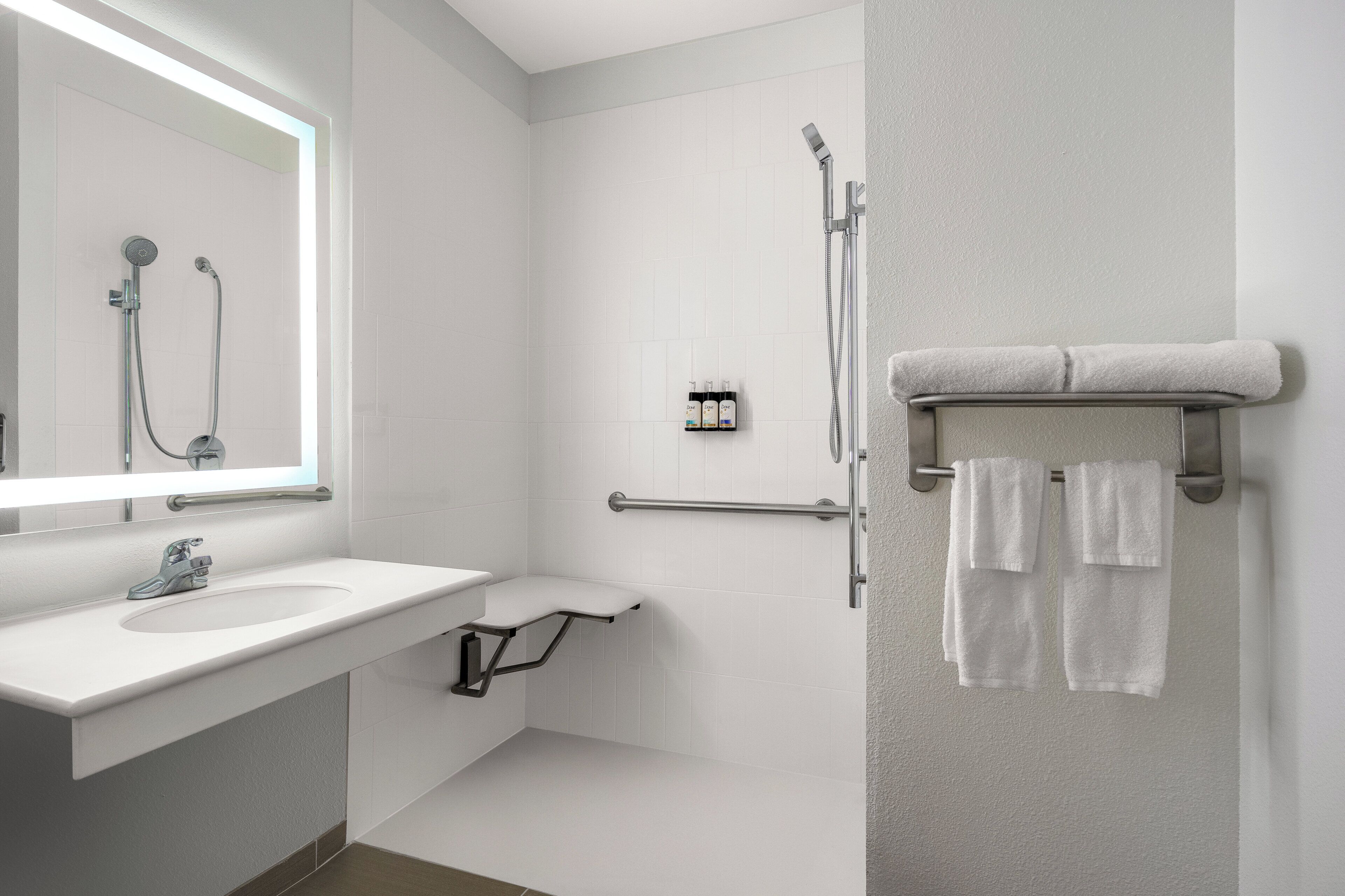 Un baño moderno con un lavamanos blanco, un espejo, una ducha con cabezal de ducha manual y un perchero con dos toallas.
