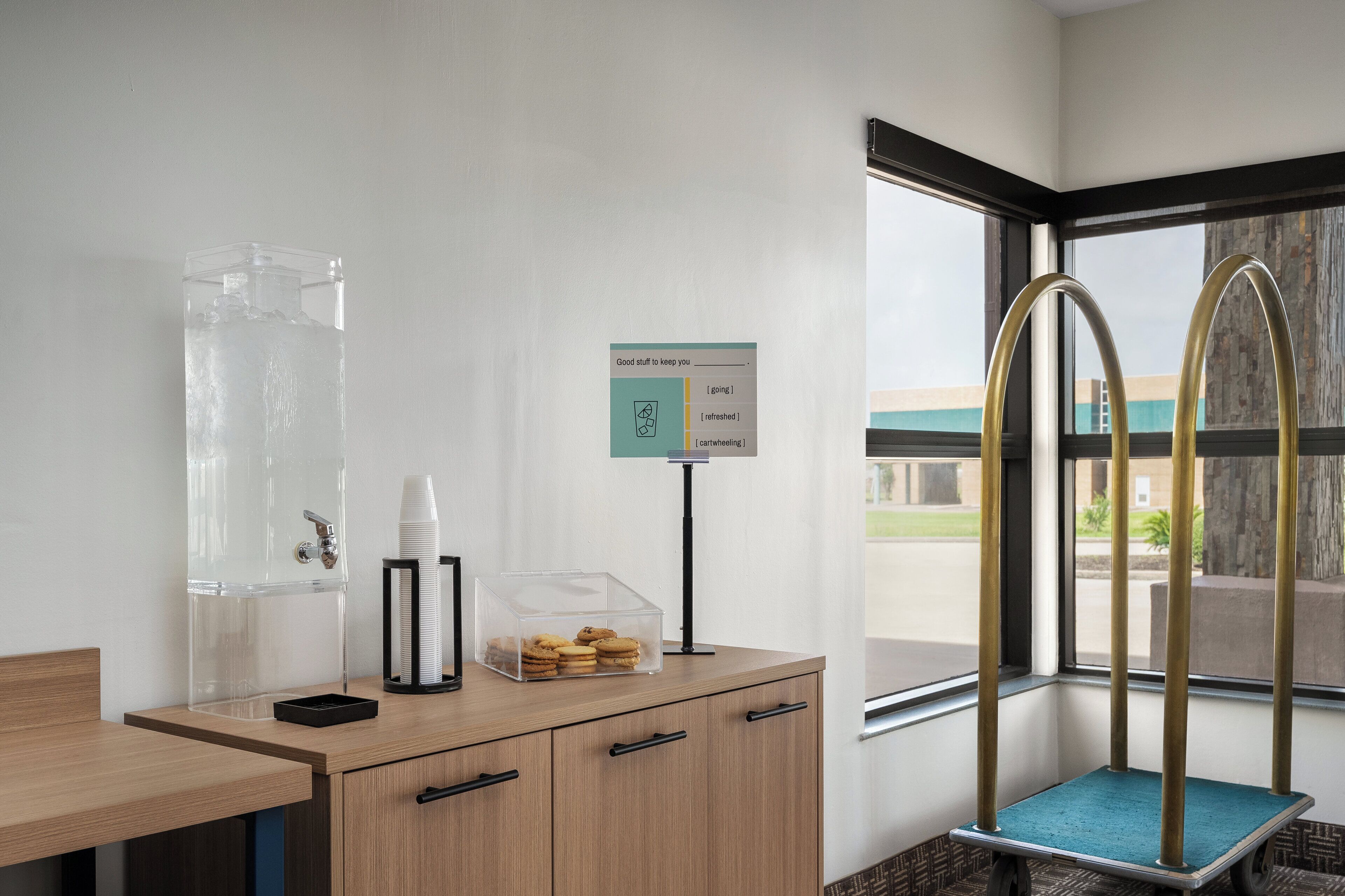 A room with a water dispenser, a sign with options, a stack of white cups, a container of cookies, and a window with a view.