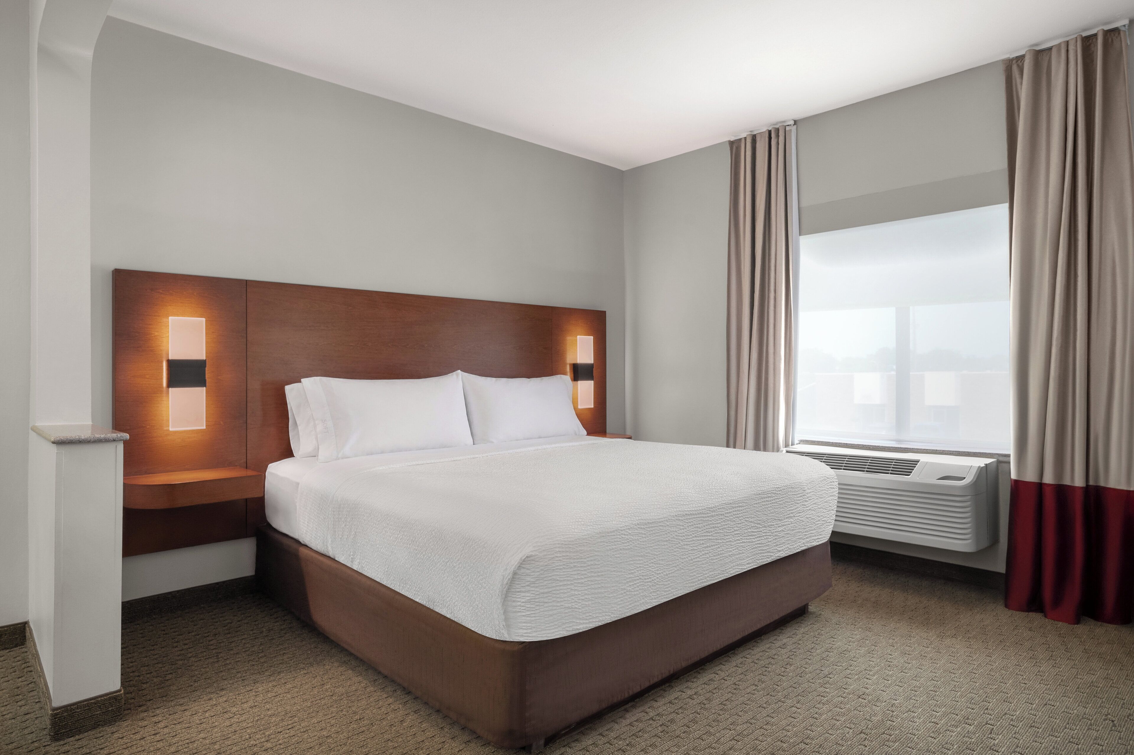 A hotel room with a large bed, a nightstand, a wall-mounted lamp, a window with curtains, and an air conditioning unit.