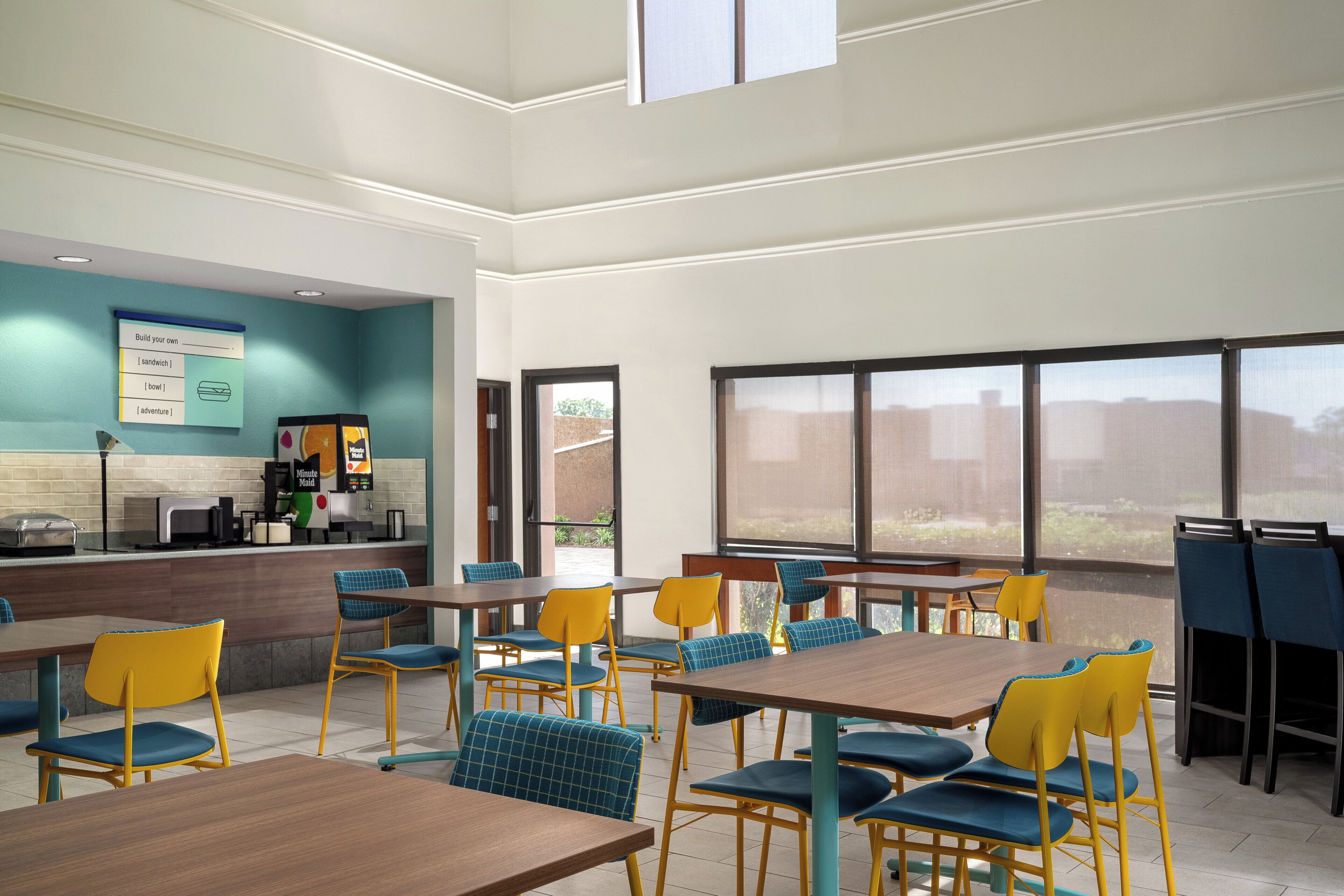 A modern cafeteria with multiple tables and chairs, a counter with a menu board, and large windows providing natural light.