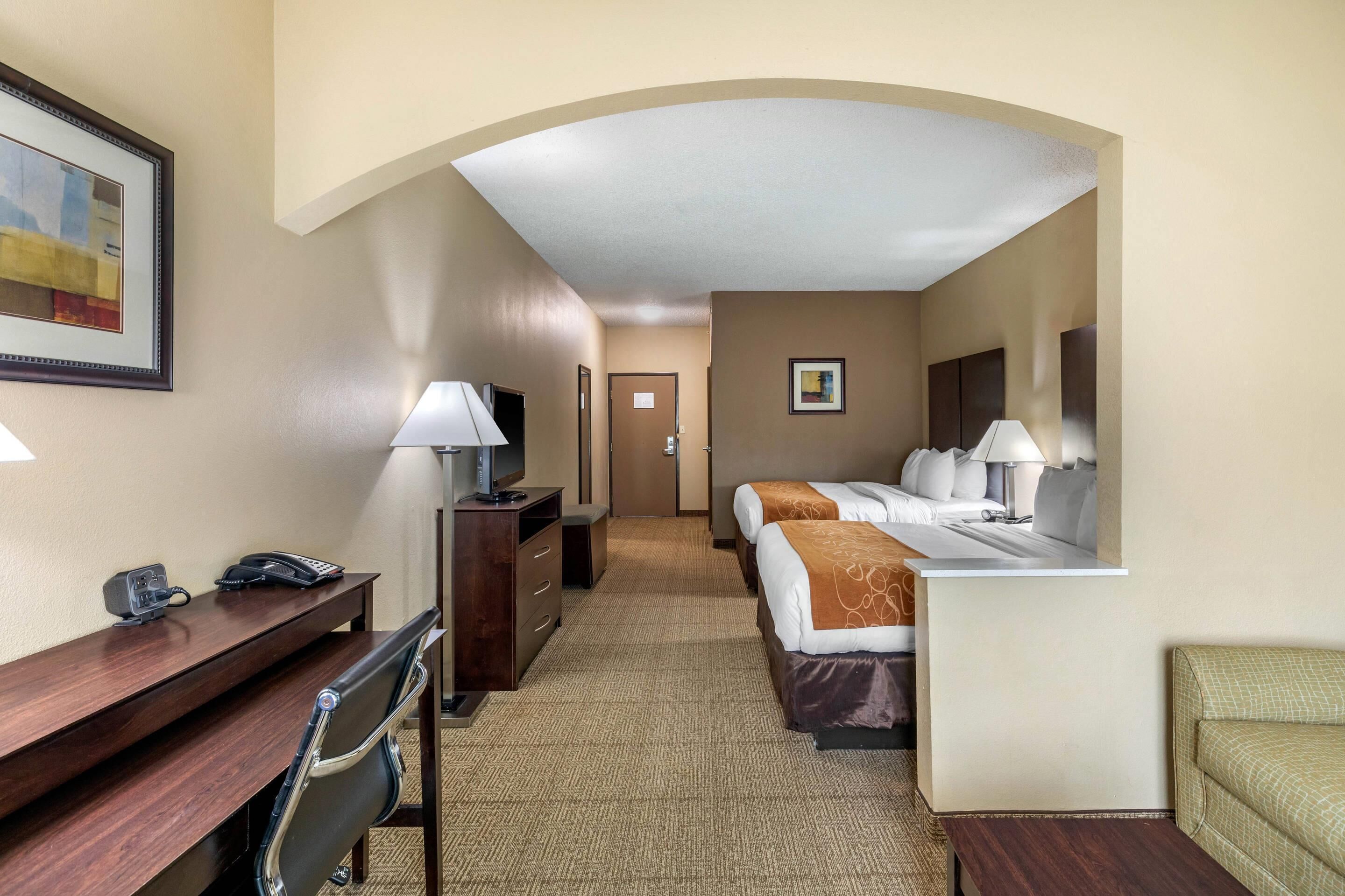 A hotel room with a desk, two beds, a chair, a TV, and a lamp.
