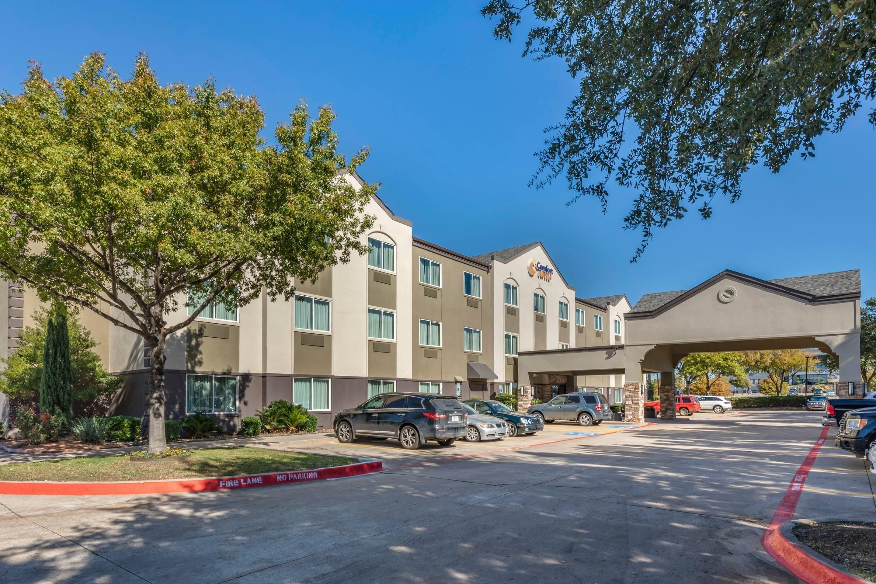 A hotel with a parking lot, a tree, and a clear blue sky.