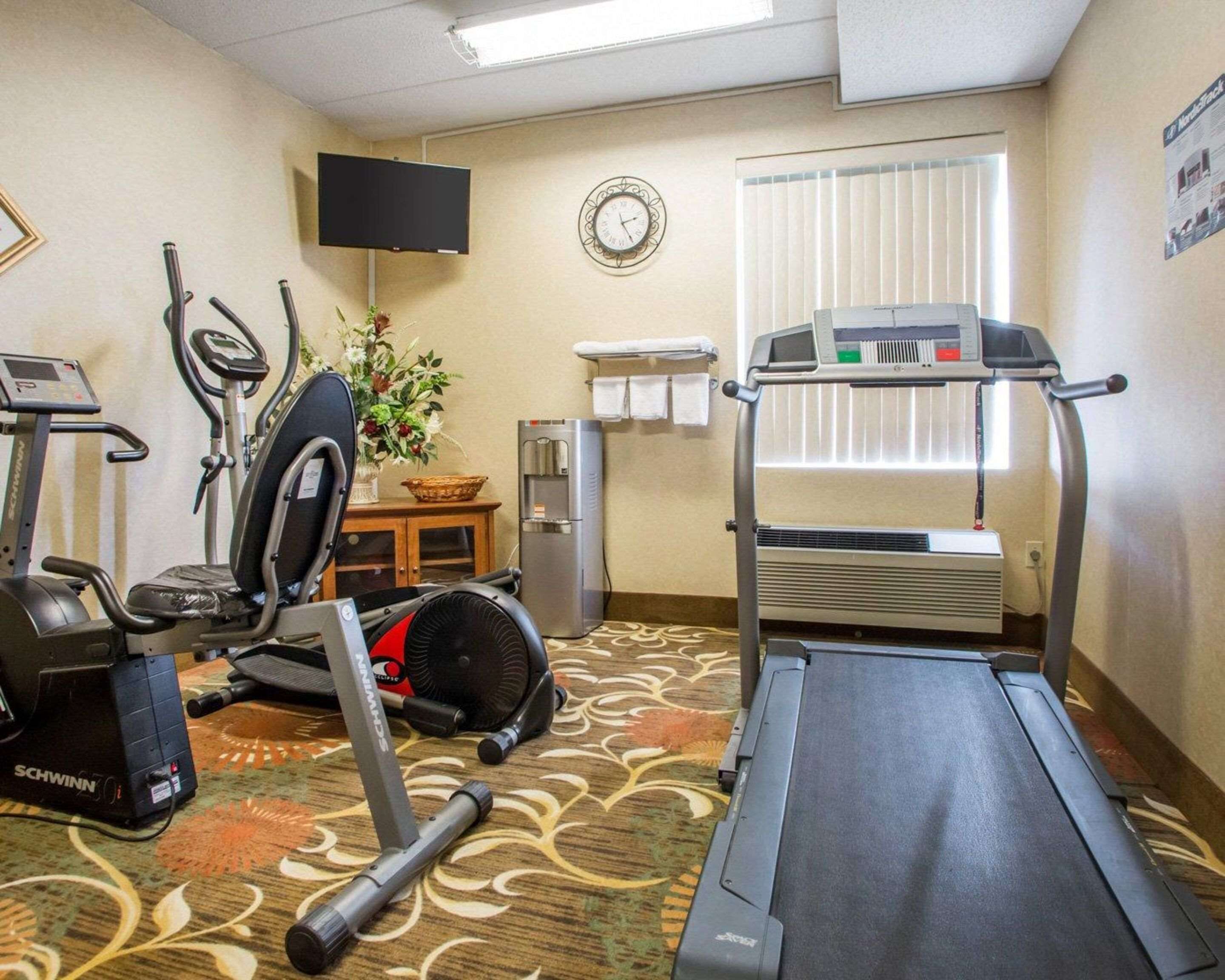 A hotel room with a treadmill, stationary bike, and elliptical machine. There is a clock on the wall, a window with blinds, and a water cooler.